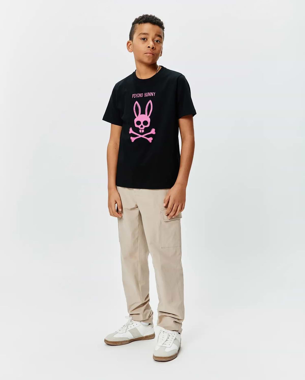 Kids Adler Graphic Tee, a black cotton jersey t-shirt with a large pink Psycho Bunny skull and crossbones logo and text on the chest, styled with beige cargo pants.