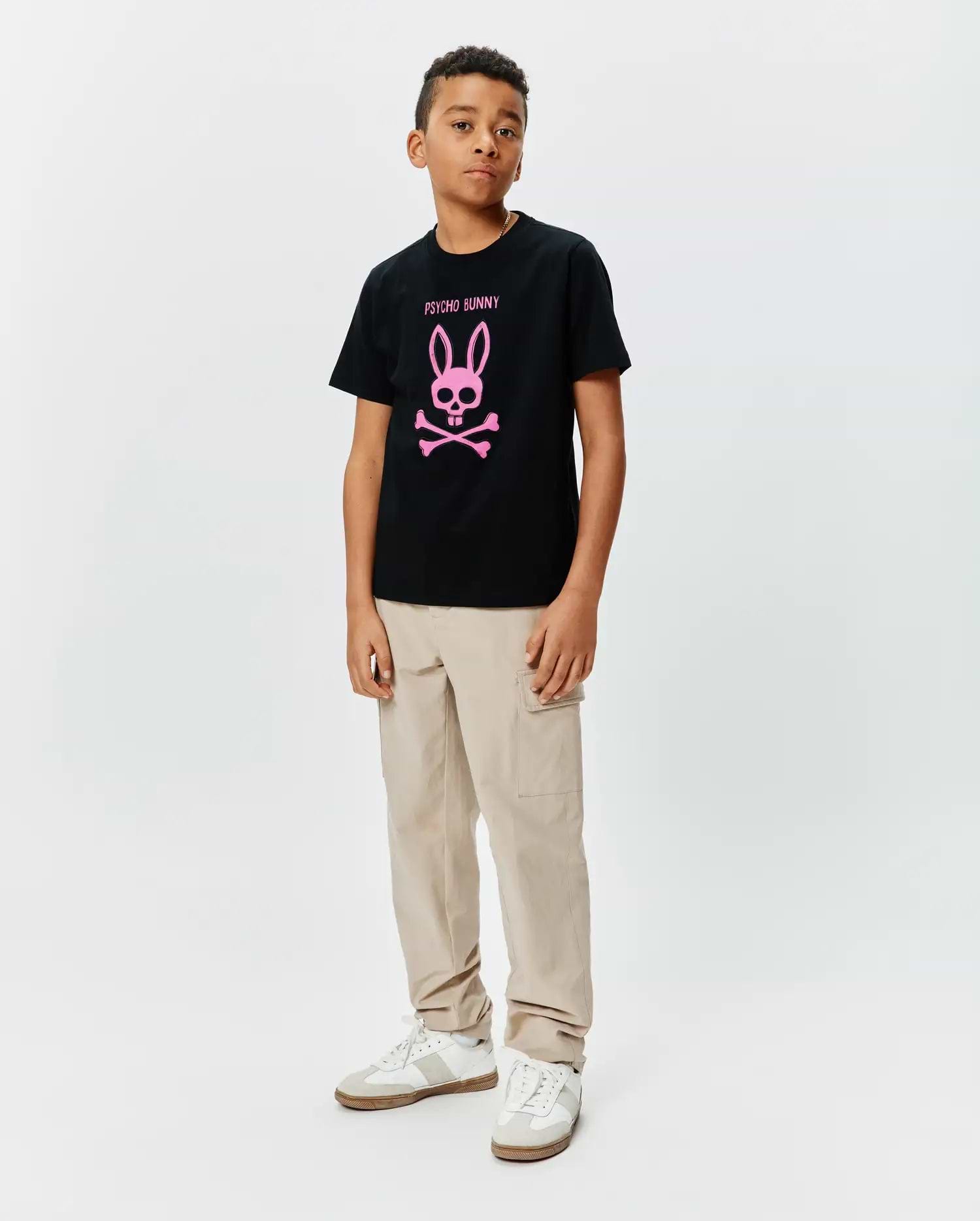 Kids Adler Graphic Tee, a black cotton jersey t-shirt with a large pink Psycho Bunny skull and crossbones logo and text on the chest, styled with beige cargo pants.