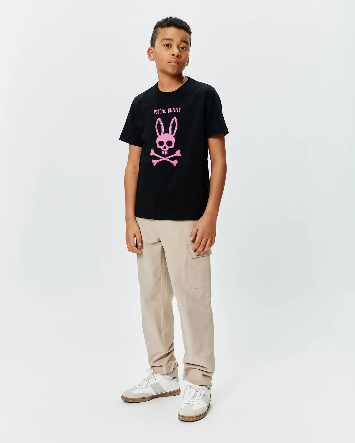 Kids Adler Graphic Tee, a black cotton jersey t-shirt with a large pink Psycho Bunny skull and crossbones logo and text on the chest, styled with beige cargo pants.