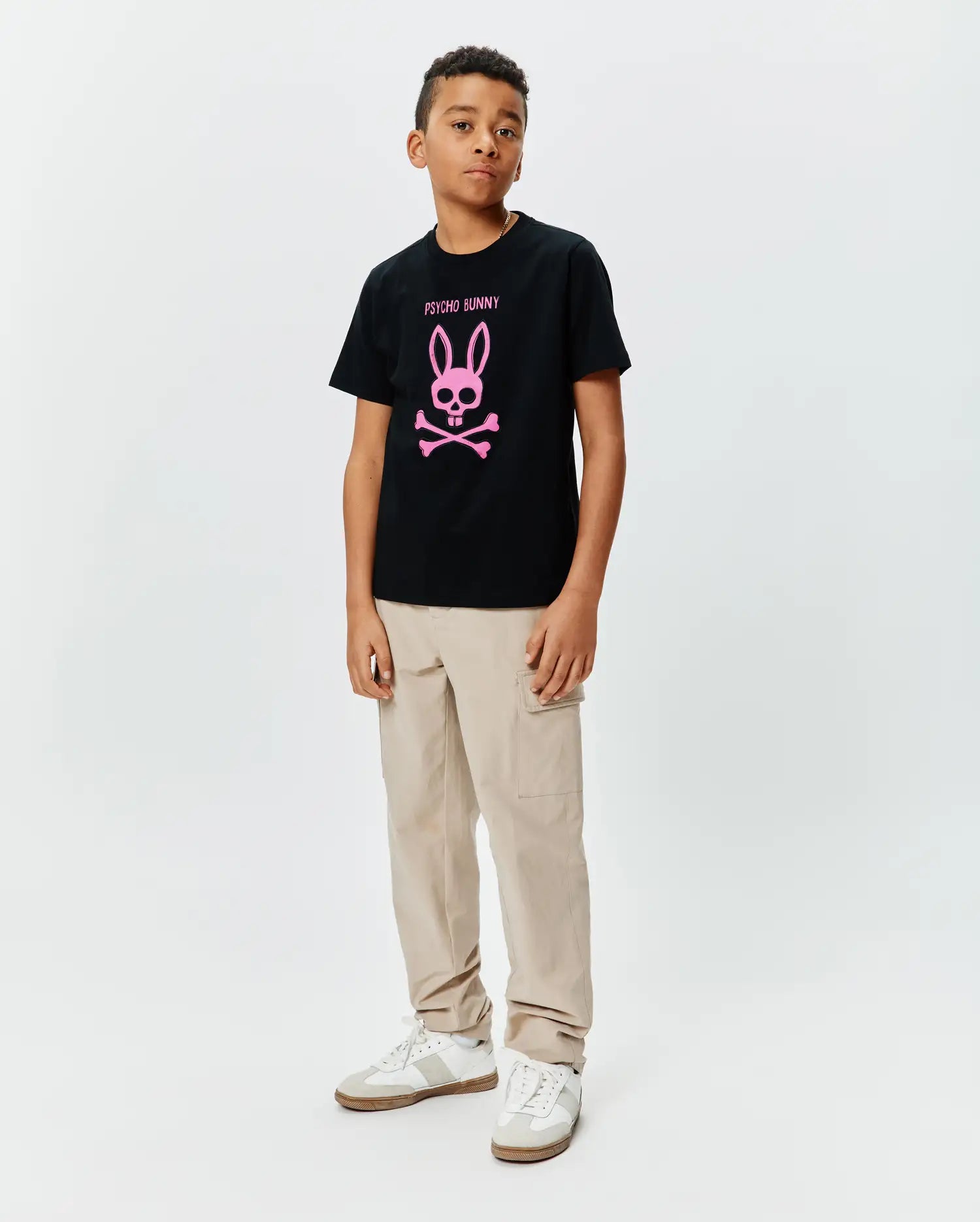 Kids Adler Graphic Tee, a black cotton jersey t-shirt with a large pink Psycho Bunny skull and crossbones logo and text on the chest, styled with beige cargo pants.