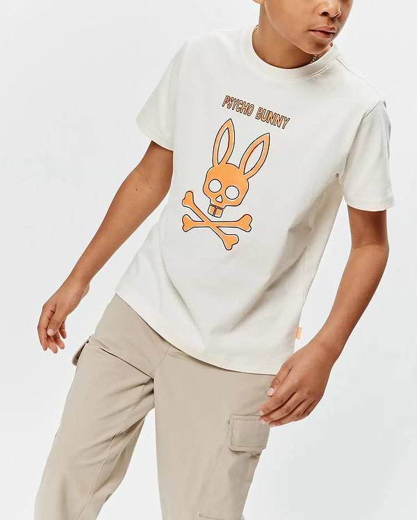 Kids Adler Graphic Tee in cream, featuring a relaxed fit and soft jersey fabric. A large orange and black outlined Psycho Bunny skull and crossbones graphic with PSYCHO BUNNY text is centered on the chest, complemented by a small orange side tag.
