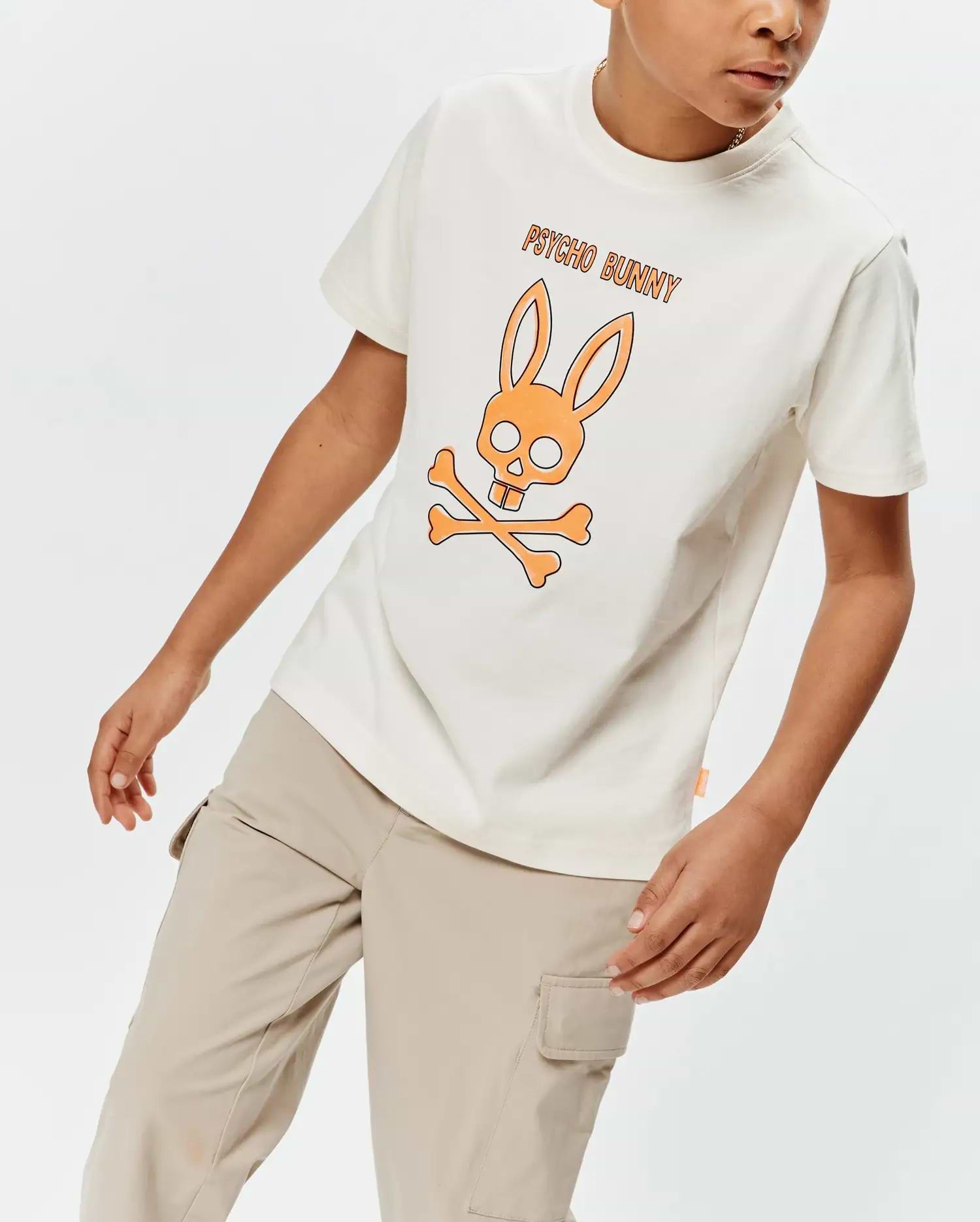 Kids Adler Graphic Tee in cream, featuring a relaxed fit and soft jersey fabric. A large orange and black outlined Psycho Bunny skull and crossbones graphic with PSYCHO BUNNY text is centered on the chest, complemented by a small orange side tag.