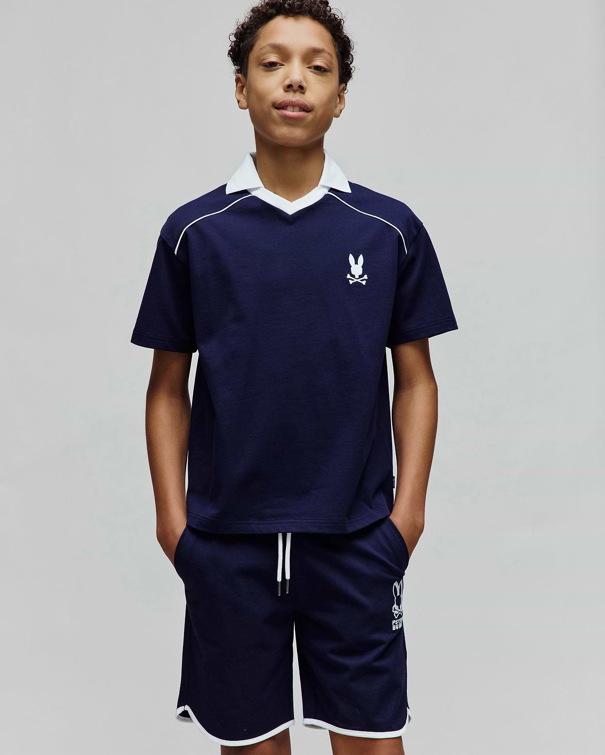 Kids Toby Jersey navy blue jersey top with white V-neck collar, white shoulder piping, and small embroidered Psycho Bunny logo. Matching navy shorts have white hem piping, drawstrings, and a larger Psycho Bunny logo.