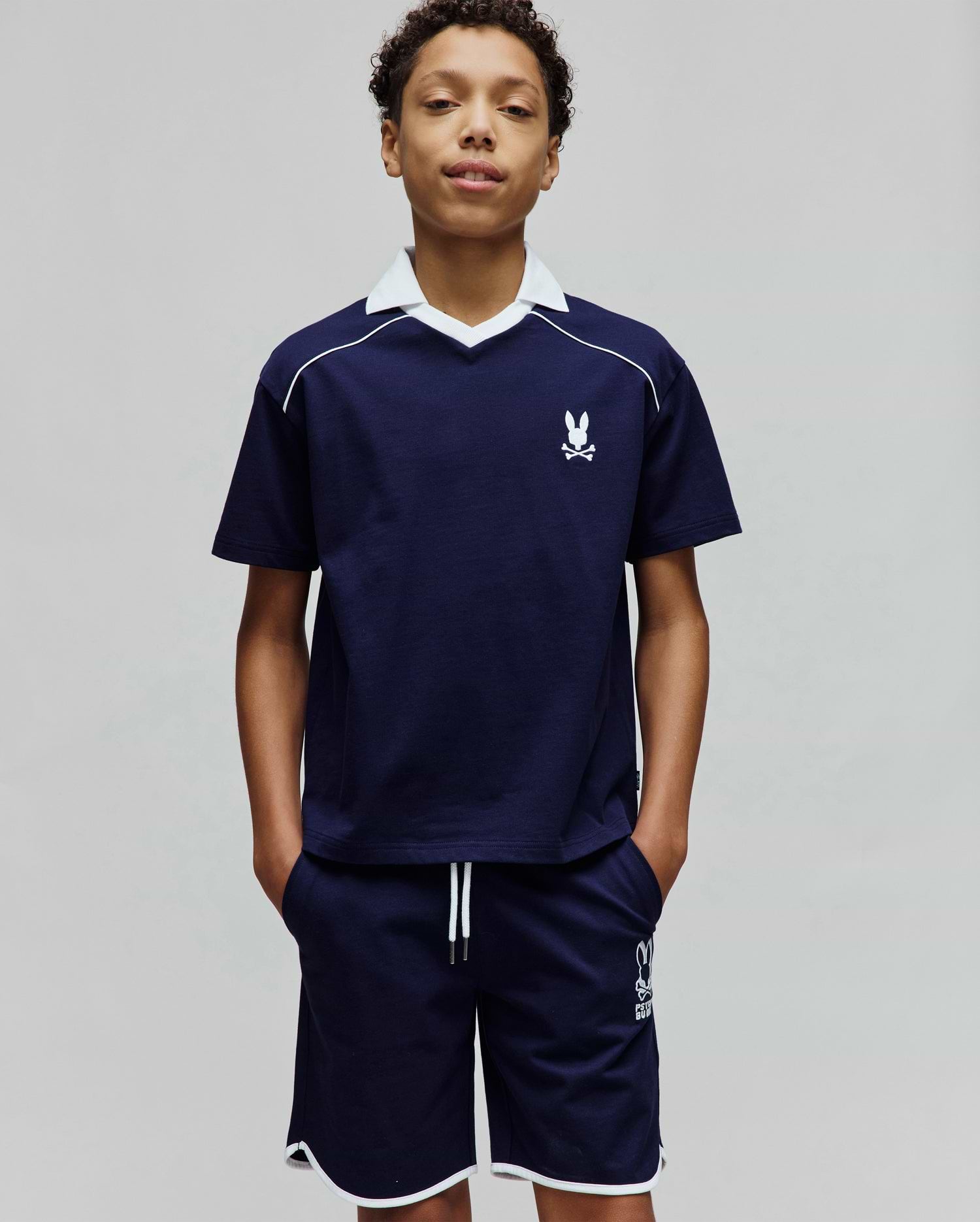 Kids Toby Jersey navy blue jersey top with white V-neck collar, white shoulder piping, and small embroidered Psycho Bunny logo. Matching navy shorts have white hem piping, drawstrings, and a larger Psycho Bunny logo.