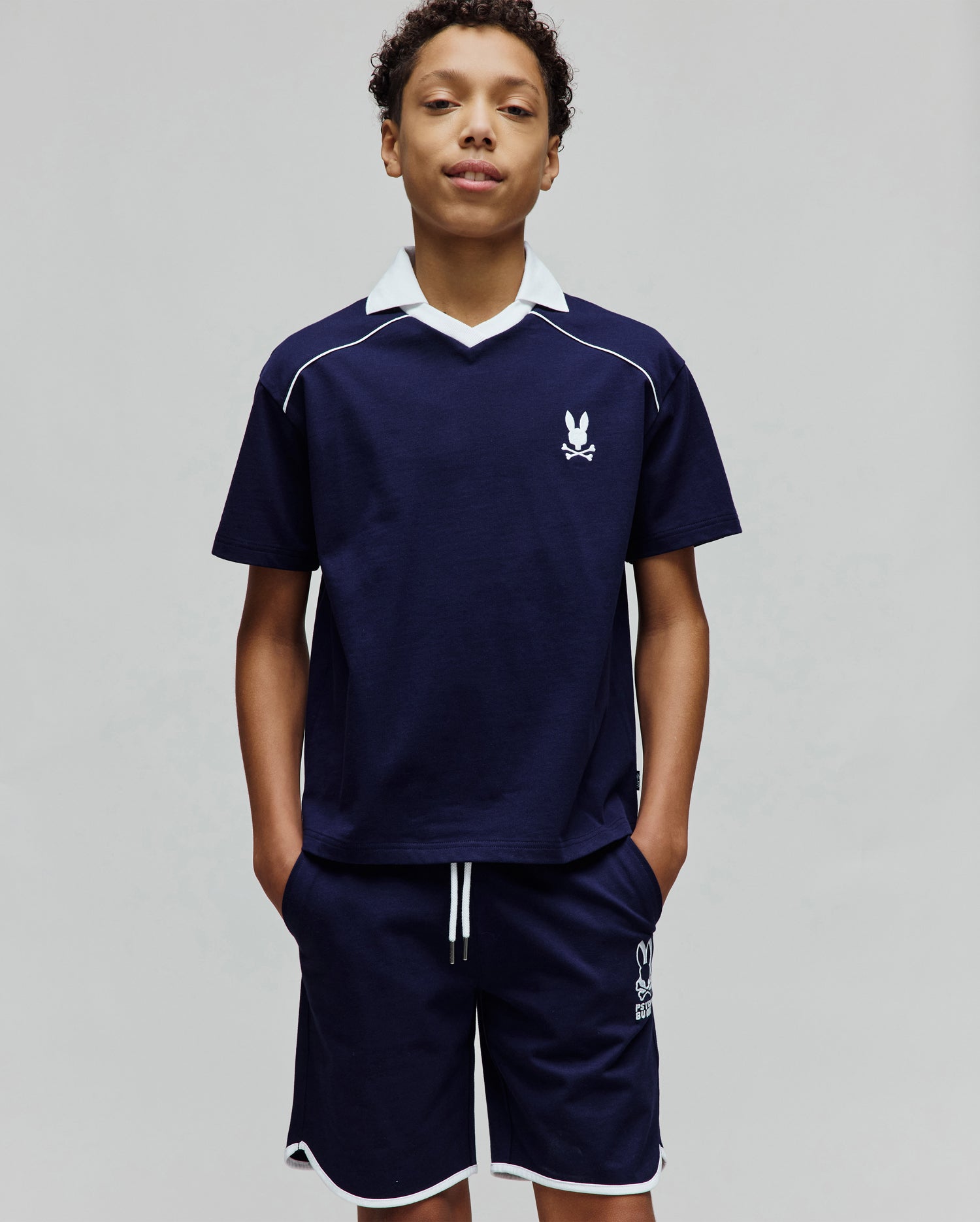 Kids Toby Jersey navy blue jersey top with white V-neck collar, white shoulder piping, and small embroidered Psycho Bunny logo. Matching navy shorts have white hem piping, drawstrings, and a larger Psycho Bunny logo.