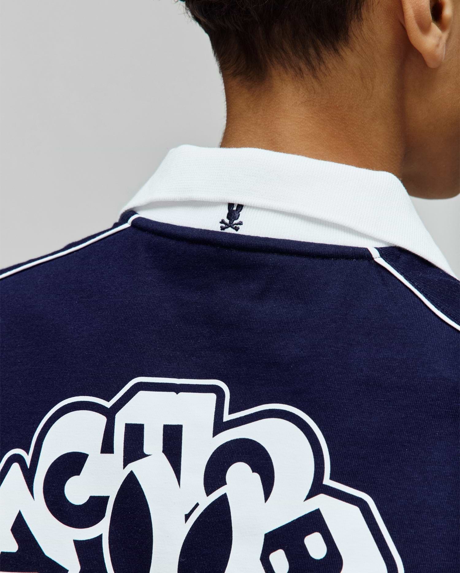 Kids Toby Jersey navy blue jersey polo, with a white ribbed collar, white shoulder piping, small embroidered Psycho Bunny skull logo on the back collar, and large white graphic print on the back.