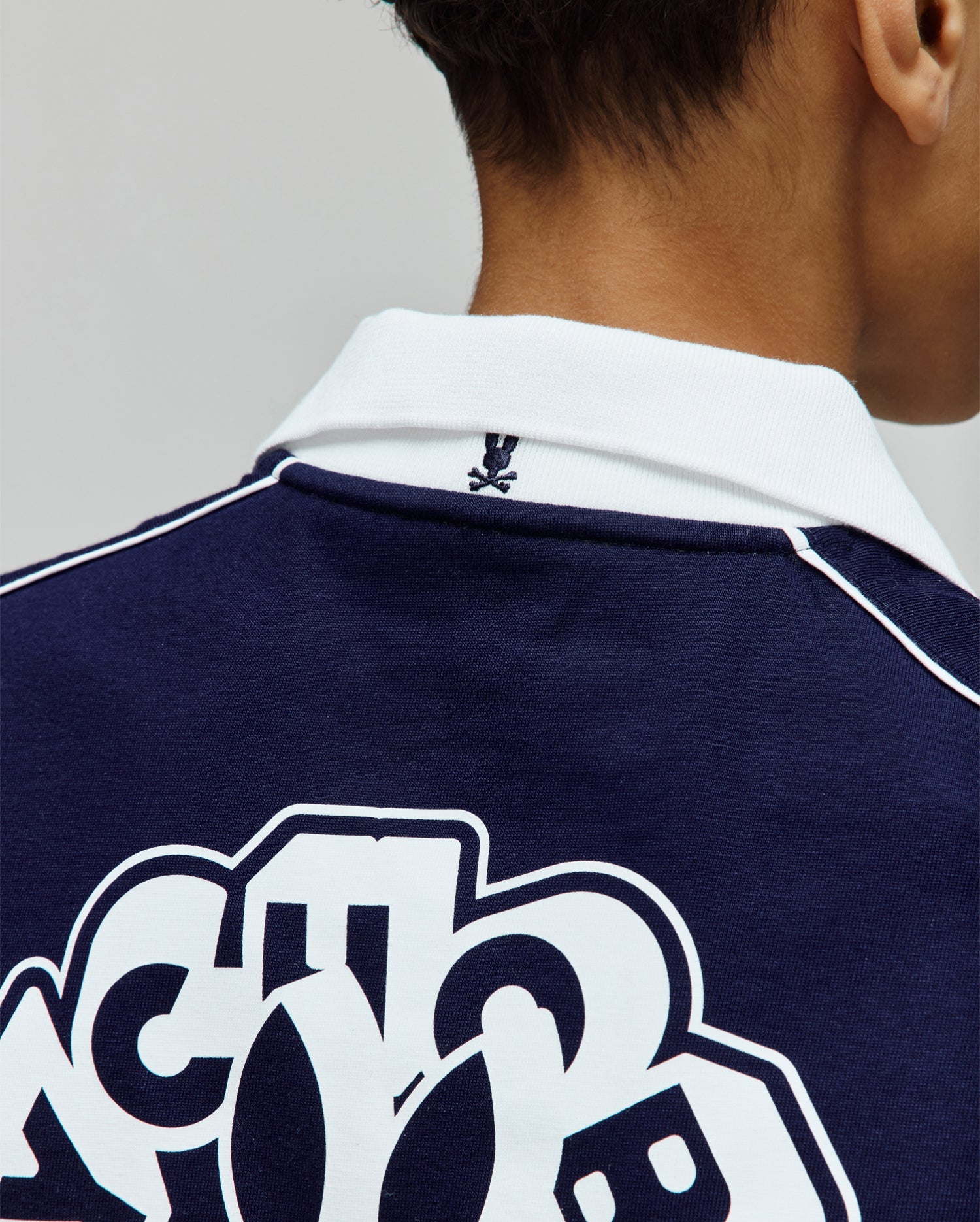 Kids Toby Jersey navy blue jersey polo, with a white ribbed collar, white shoulder piping, small embroidered Psycho Bunny skull logo on the back collar, and large white graphic print on the back.
