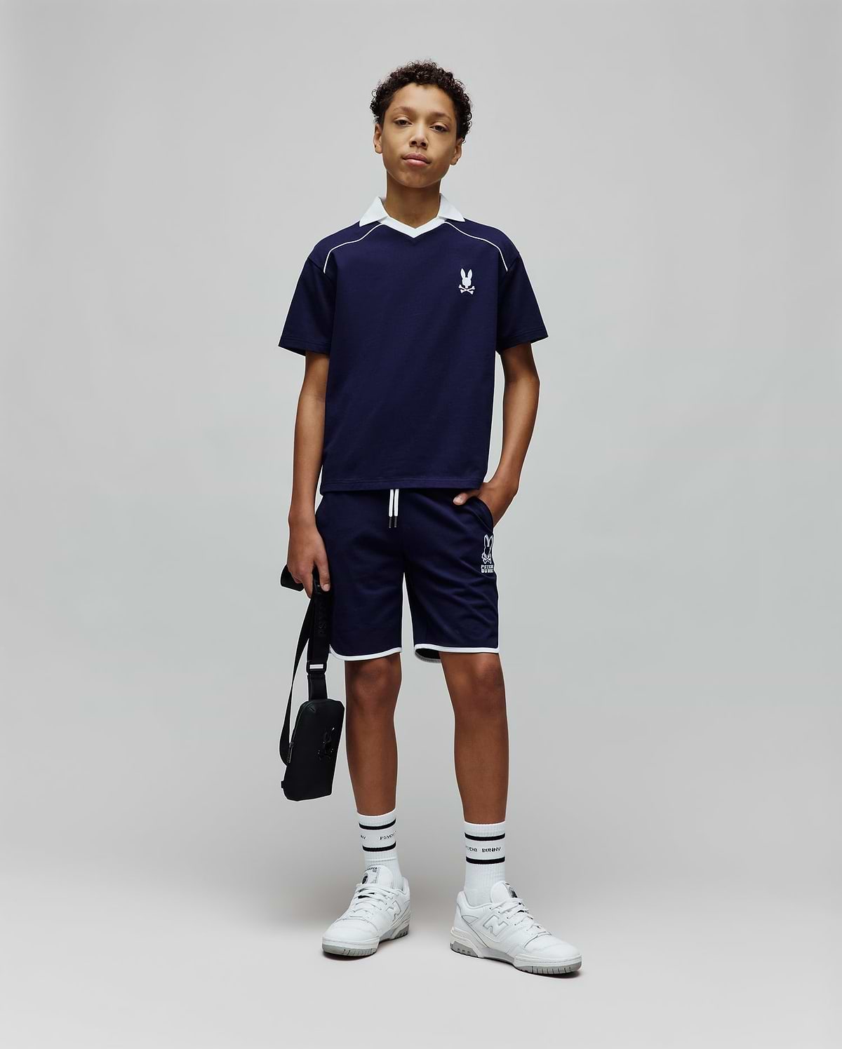 Navy blue Kids Toby Jersey t-shirt with white V-neck collar, shoulder stripe, and small embroidered Psycho Bunny skull logo on the left chest, paired with matching shorts featuring white piping and a large logo print.
