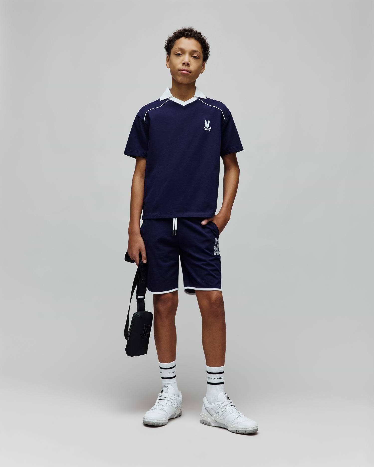Navy blue Kids Toby Jersey t-shirt with white V-neck collar, shoulder stripe, and small embroidered Psycho Bunny skull logo on the left chest, paired with matching shorts featuring white piping and a large logo print.