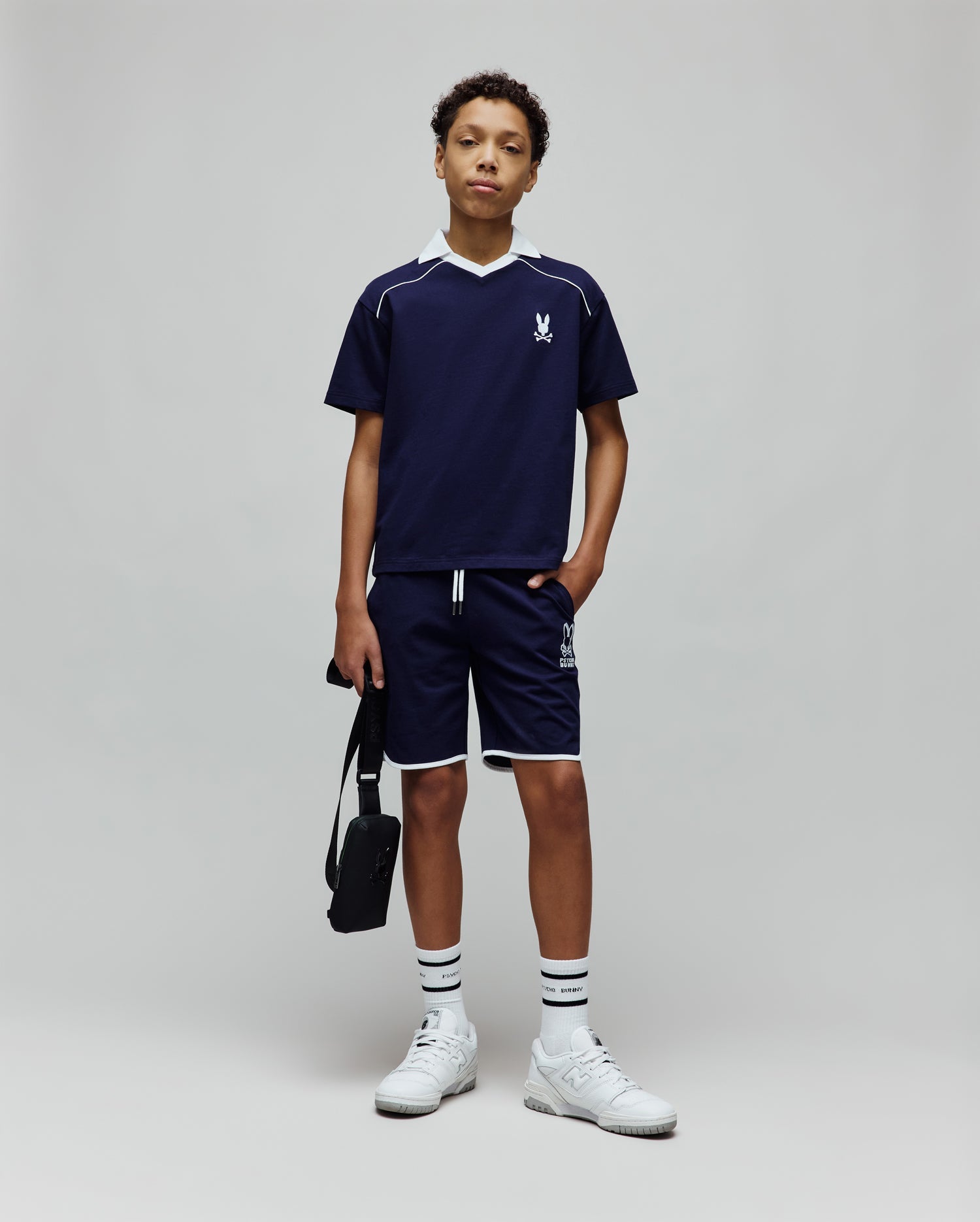 Navy blue Kids Toby Jersey t-shirt with white V-neck collar, shoulder stripe, and small embroidered Psycho Bunny skull logo on the left chest, paired with matching shorts featuring white piping and a large logo print.