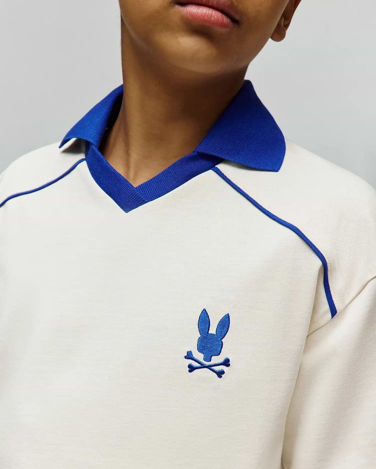 Kids Toby Jersey, cream jersey knit polo with royal blue ribbed collar, V-neck trim, raglan piping, and small embroidered royal blue Psycho Bunny skull logo on left chest.