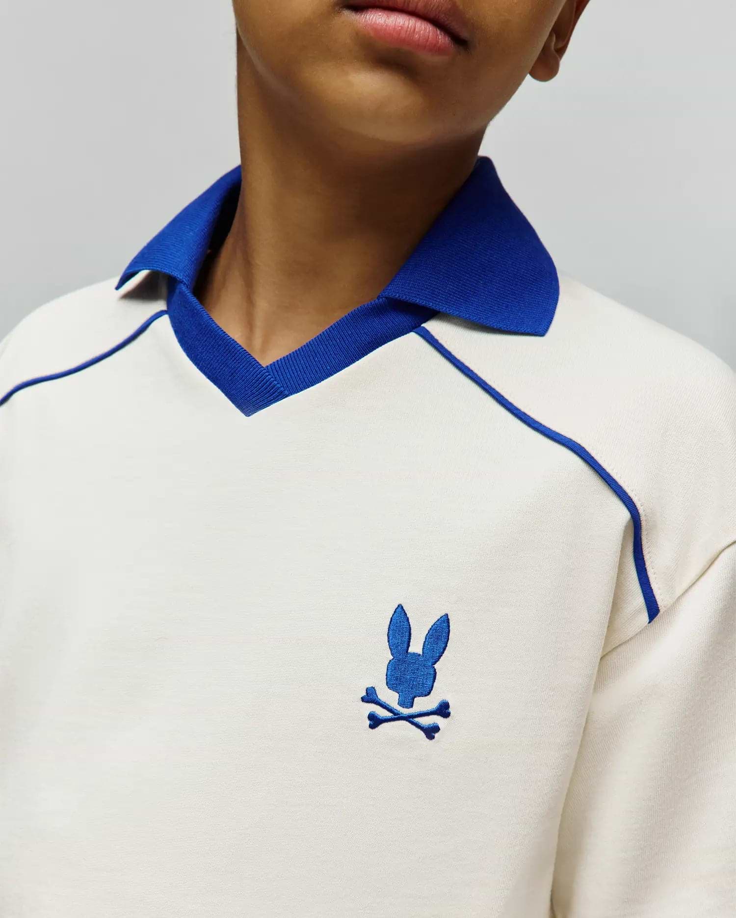 Kids Toby Jersey, cream jersey knit polo with royal blue ribbed collar, V-neck trim, raglan piping, and small embroidered royal blue Psycho Bunny skull logo on left chest.