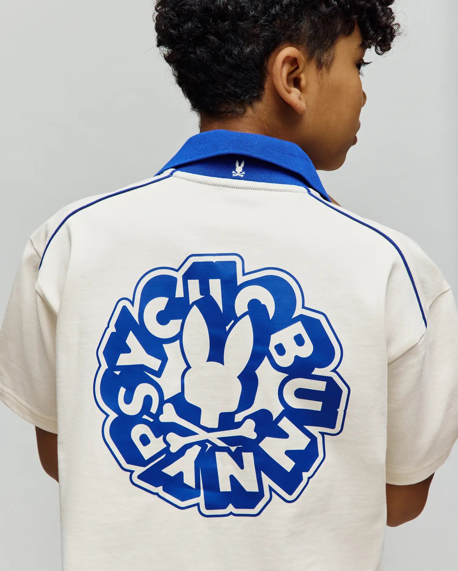 Kids Toby Jersey relaxed fit cream polo shirt with royal blue ribbed collar, shoulder piping, and a large back graphic featuring the Psycho Bunny skull and text. Small embroidered bunny skull on collar.