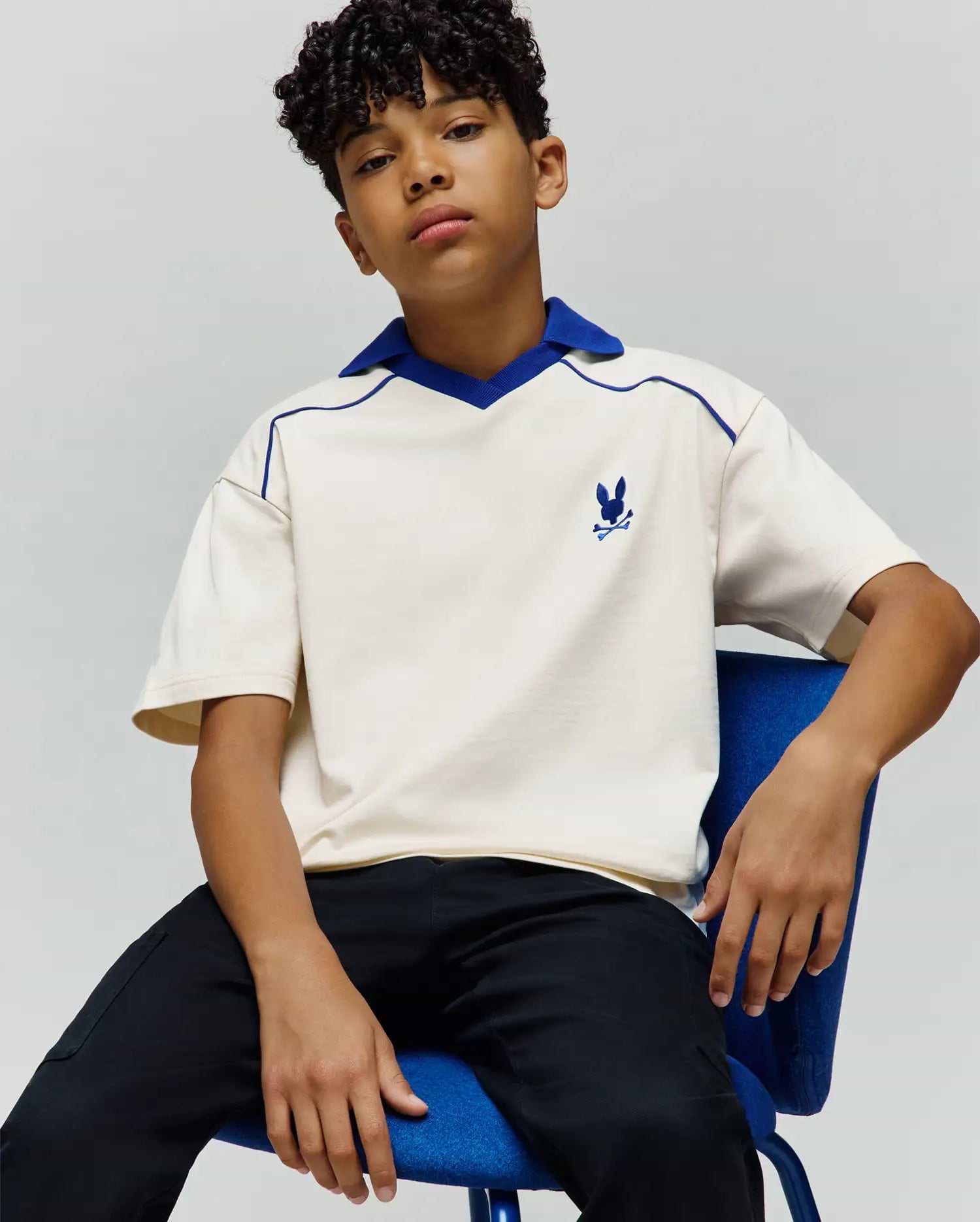 Kids Toby Jersey, a relaxed fit cream jersey polo with royal blue V-neck collar and piping, featuring a small embroidered blue Psycho Bunny skull logo on the left chest.