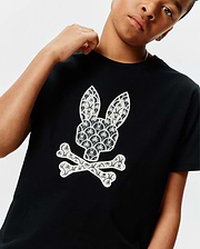 Black Kids Brad Graphic Tee, classic fit jersey, featuring a large white Psycho Bunny skull logo on the chest, intricately patterned with palm trees and mini bunny skulls.