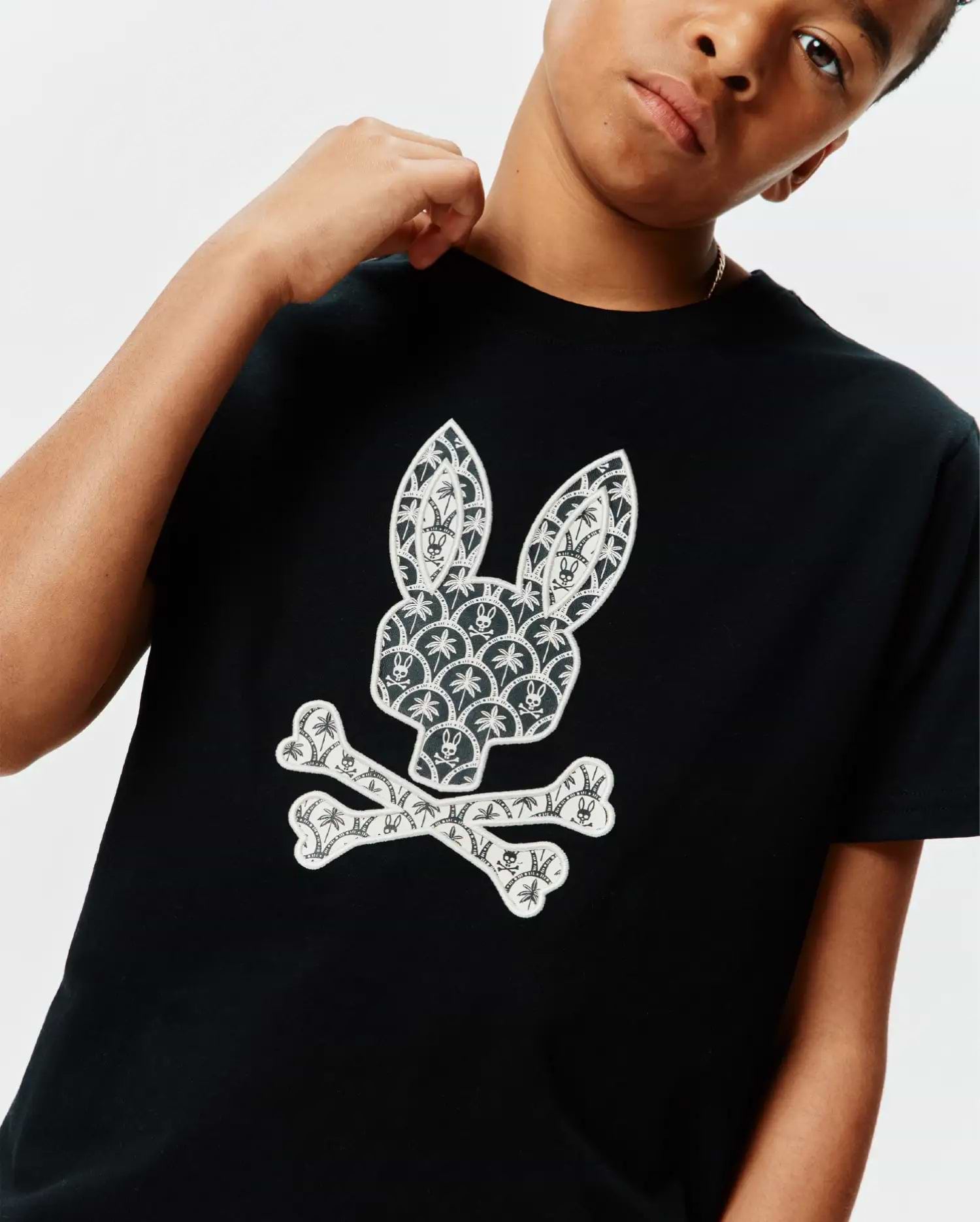 Black Kids Brad Graphic Tee, classic fit jersey, featuring a large white Psycho Bunny skull logo on the chest, intricately patterned with palm trees and mini bunny skulls.