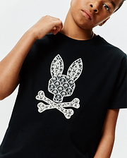 Black Kids Brad Graphic Tee, classic fit jersey, featuring a large white Psycho Bunny skull logo on the chest, intricately patterned with palm trees and mini bunny skulls.