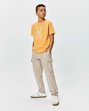 Kids Brad Graphic Tee. Mustard yellow classic fit jersey t-shirt with a large white Psycho Bunny skull and crossbones logo on the chest, featuring an intricate pattern.
