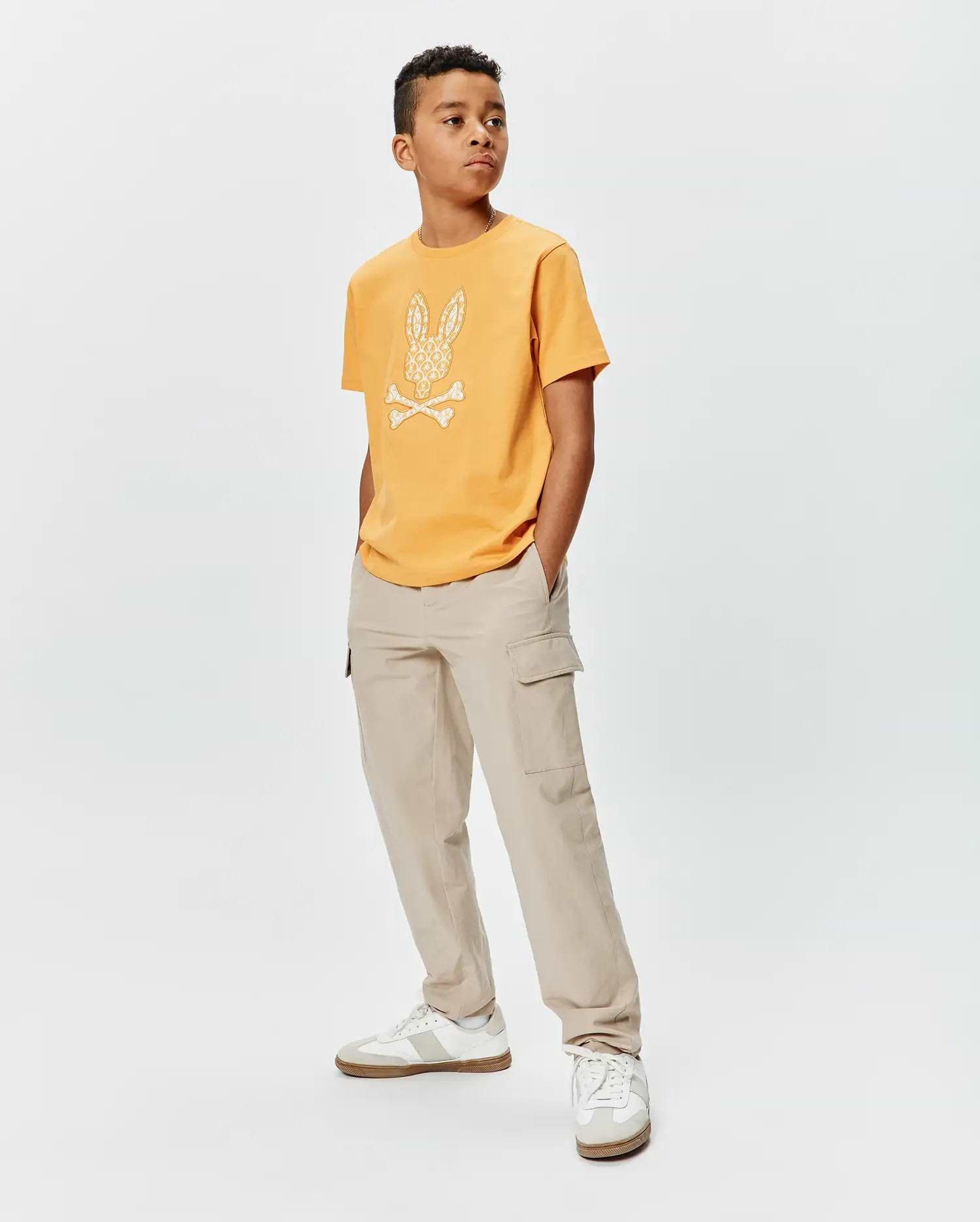 Kids Brad Graphic Tee. Mustard yellow classic fit jersey t-shirt with a large white Psycho Bunny skull and crossbones logo on the chest, featuring an intricate pattern.