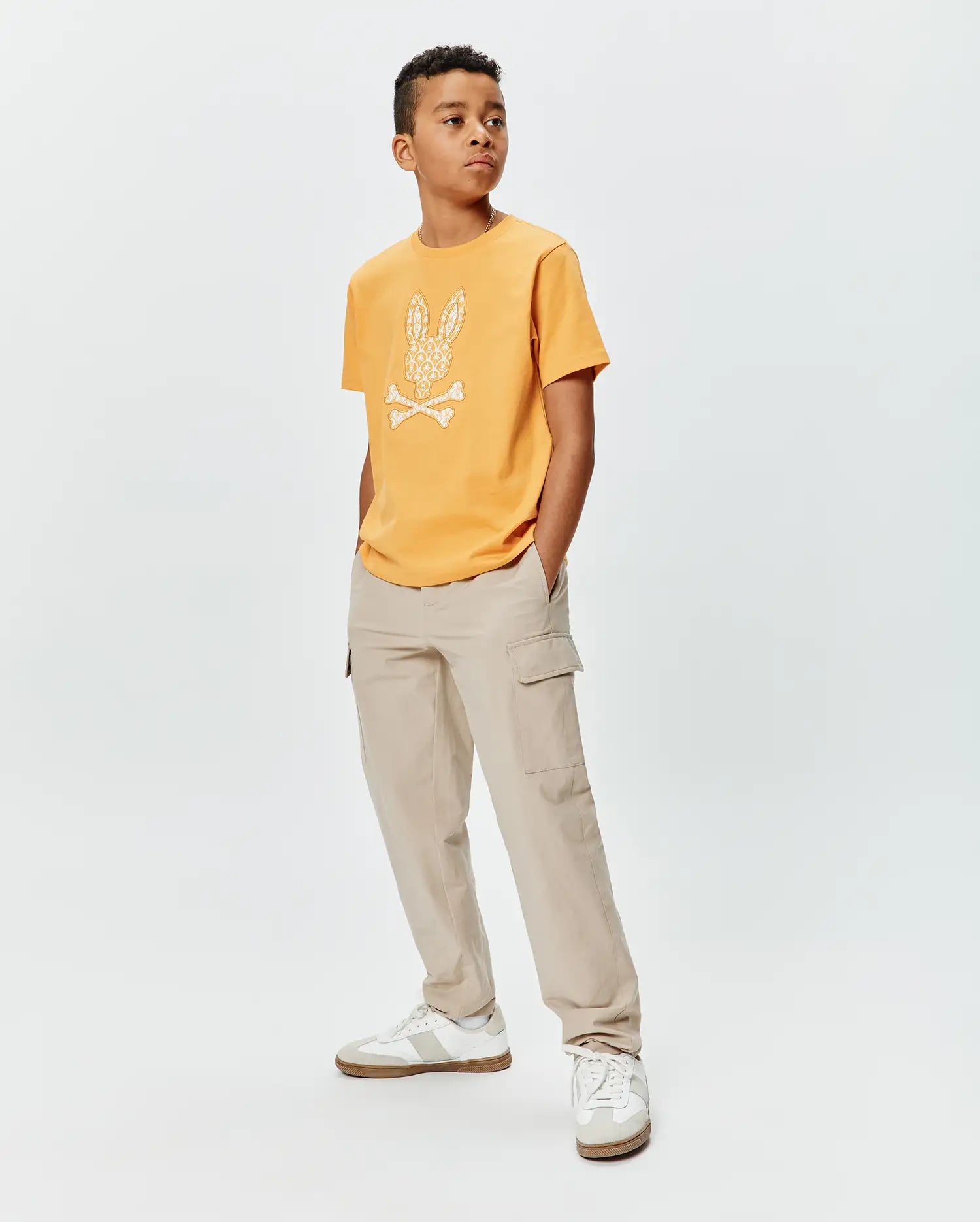 Kids Brad Graphic Tee. Mustard yellow classic fit jersey t-shirt with a large white Psycho Bunny skull and crossbones logo on the chest, featuring an intricate pattern.