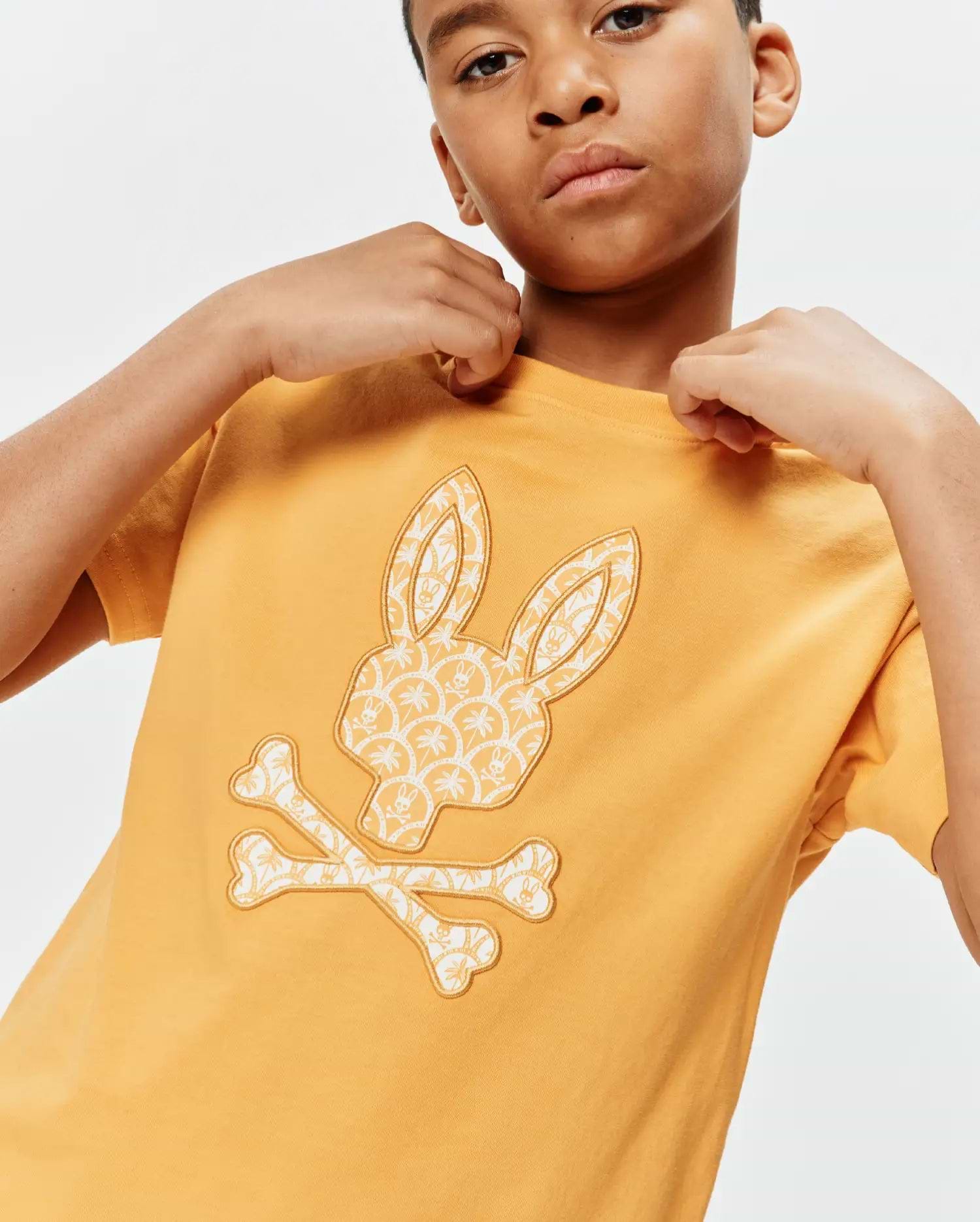 Kids Brad Graphic Tee in mustard yellow, featuring a large central Psycho Bunny skull and crossbones logo filled with a cream palm tree and mini bunny skull pattern, classic fit jersey knit.
