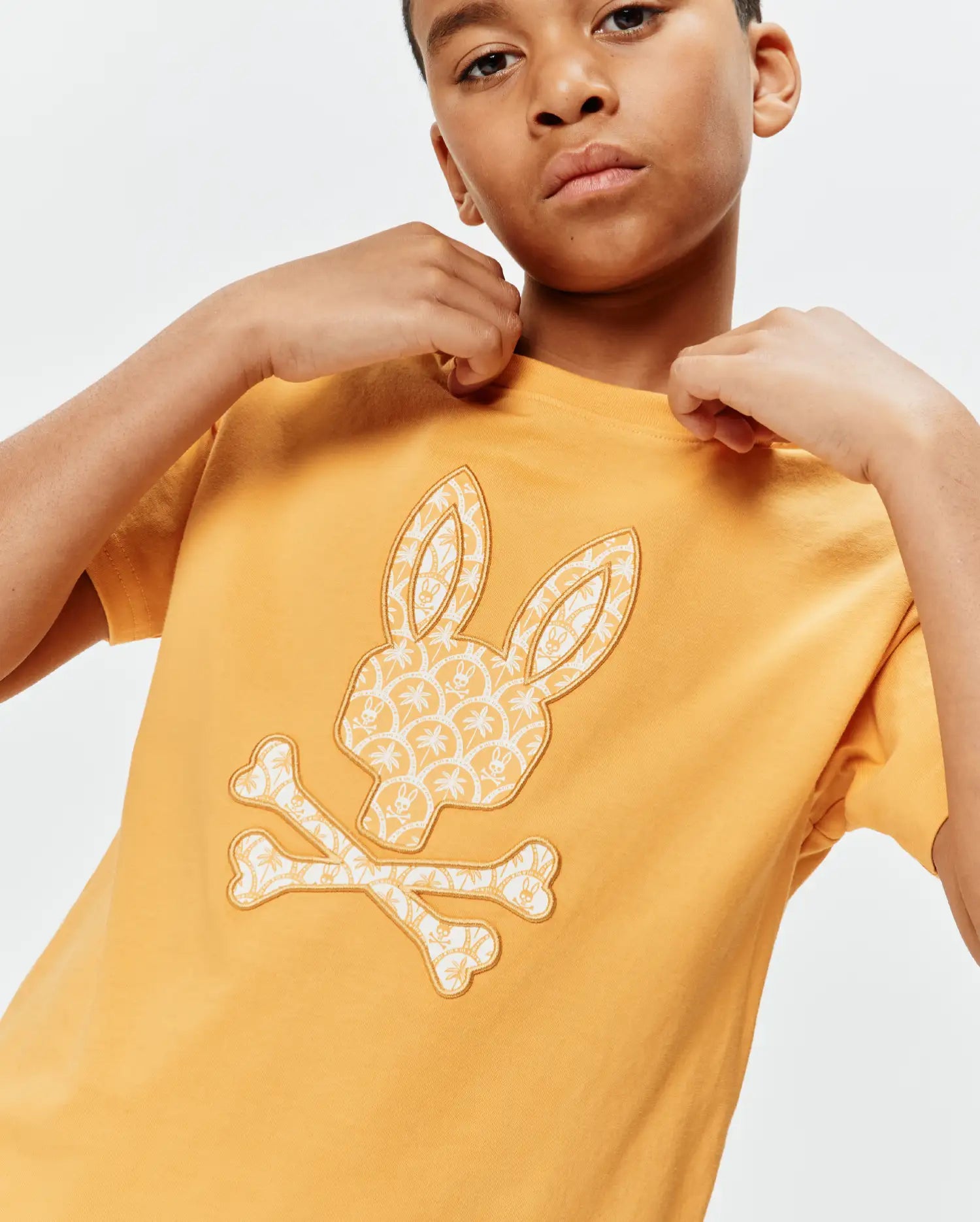 Kids Brad Graphic Tee in mustard yellow, featuring a large central Psycho Bunny skull and crossbones logo filled with a cream palm tree and mini bunny skull pattern, classic fit jersey knit.