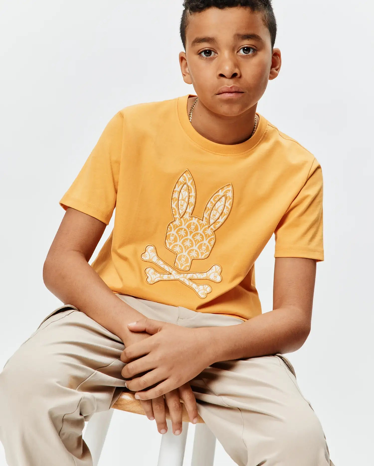 Kids Brad Graphic Tee, a classic fit mustard yellow jersey t-shirt featuring a large central white Psycho Bunny skull and crossbones graphic with an intricate pattern.