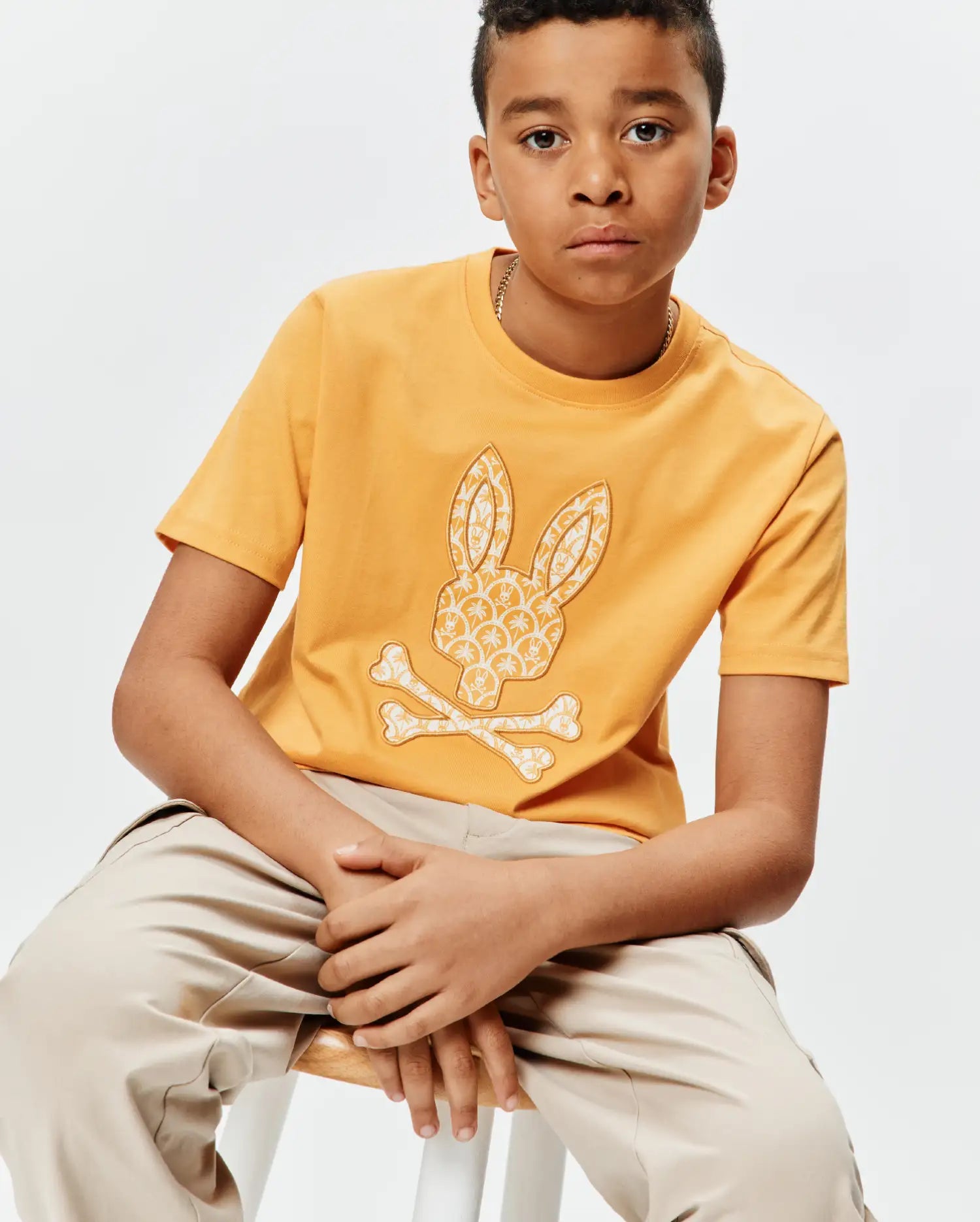 Kids Brad Graphic Tee, a classic fit mustard yellow jersey t-shirt featuring a large central white Psycho Bunny skull and crossbones graphic with an intricate pattern.