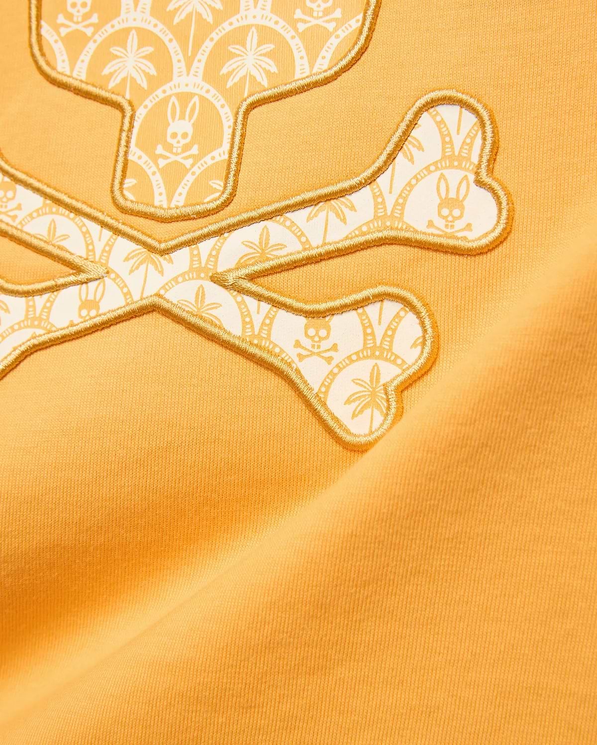 Kids Brad Graphic Tee in marigold jersey features a large gold embroidered Psycho Bunny skull and crossbones logo with a white and light orange palm tree and mini bunny skull pattern.