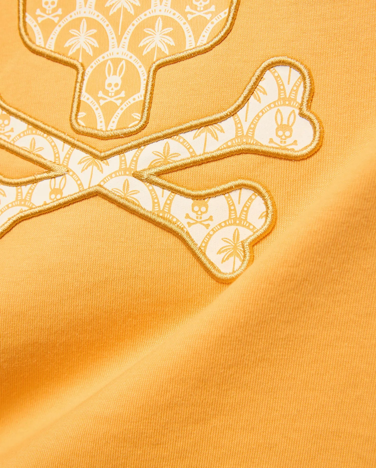 Kids Brad Graphic Tee in marigold jersey features a large gold embroidered Psycho Bunny skull and crossbones logo with a white and light orange palm tree and mini bunny skull pattern.