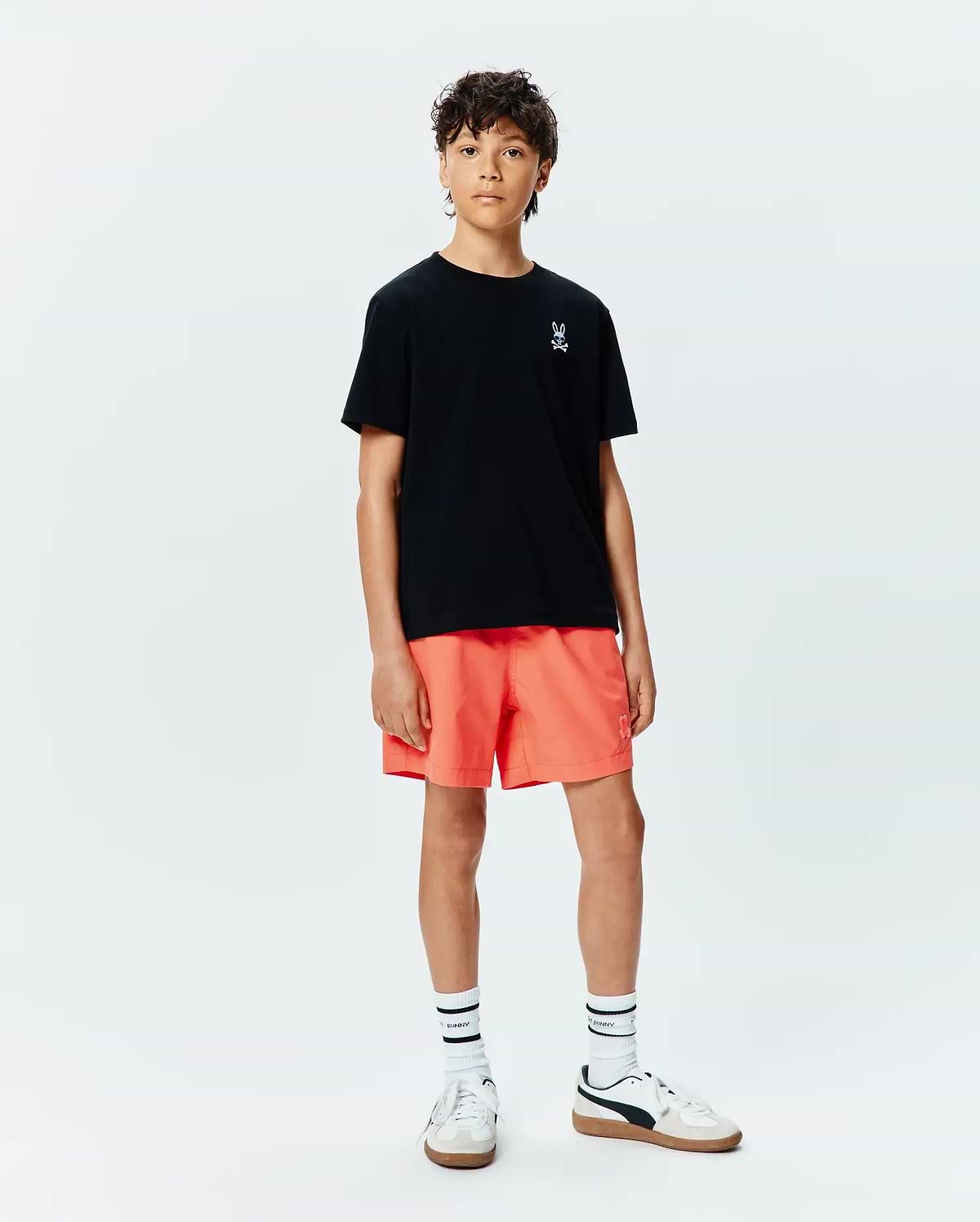 Kids Sunny Chest Graphic Tee, a black jersey t-shirt with a small white embroidered Psycho Bunny skull logo on the left chest, paired with vibrant orange relaxed-fit shorts featuring a tonal bunny logo.