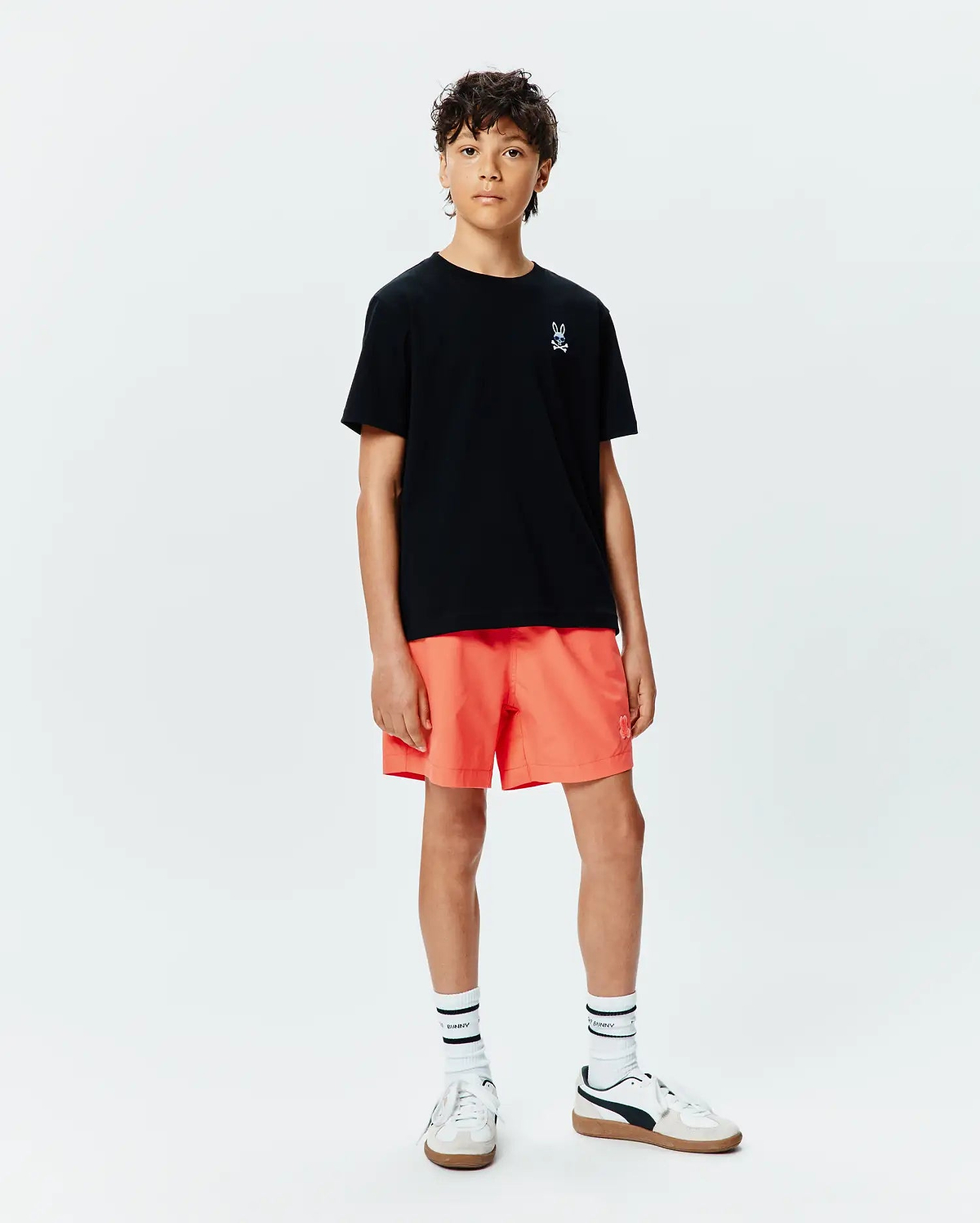 Kids Sunny Chest Graphic Tee, a black jersey t-shirt with a small white embroidered Psycho Bunny skull logo on the left chest, paired with vibrant orange relaxed-fit shorts featuring a tonal bunny logo.