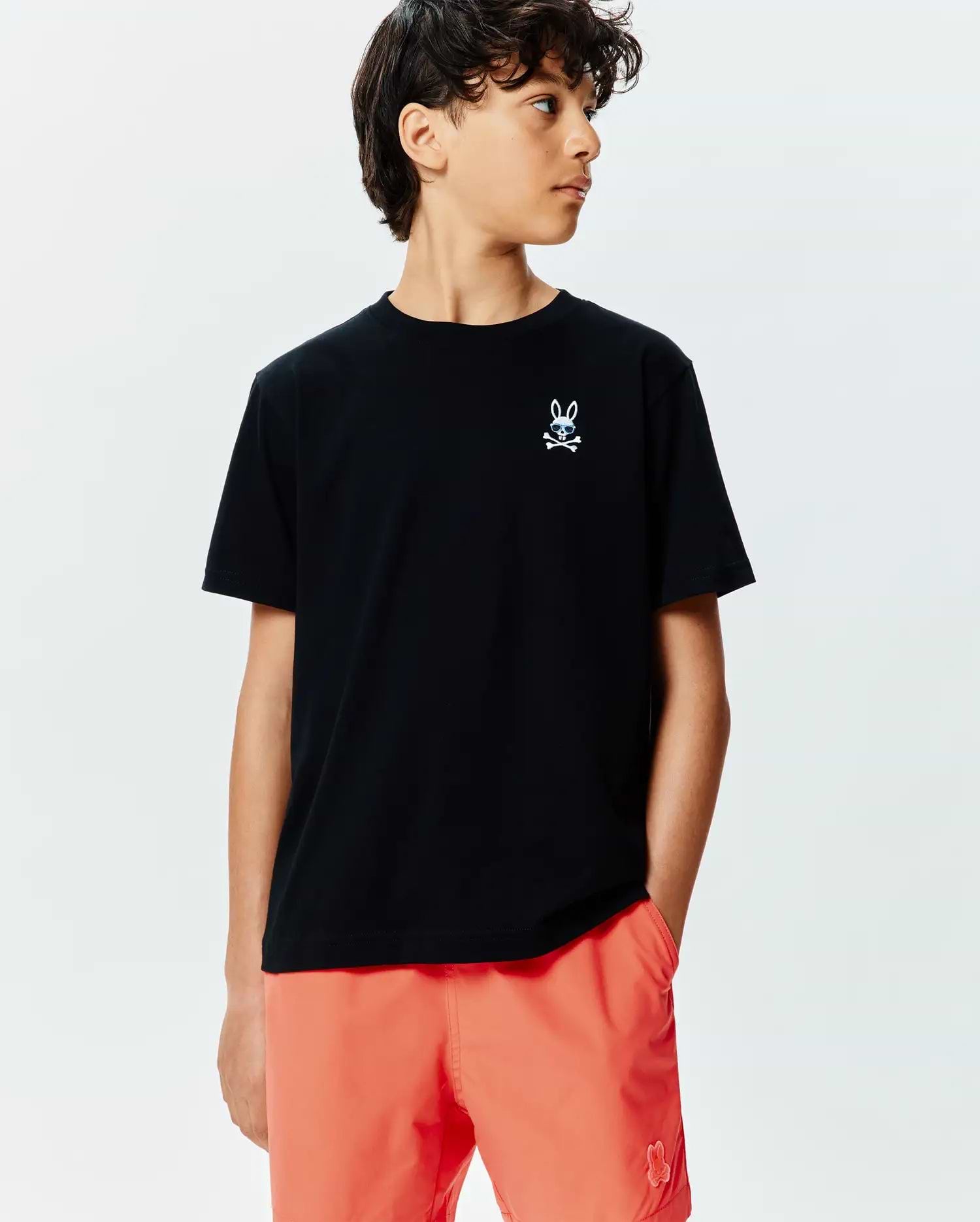 Kids Sunny Chest Graphic Tee, black classic fit jersey t-shirt, small blue/white gradient embroidered Psycho Bunny skull logo on chest. Paired with vibrant orange relaxed fit swim shorts, tonal bunny logo.