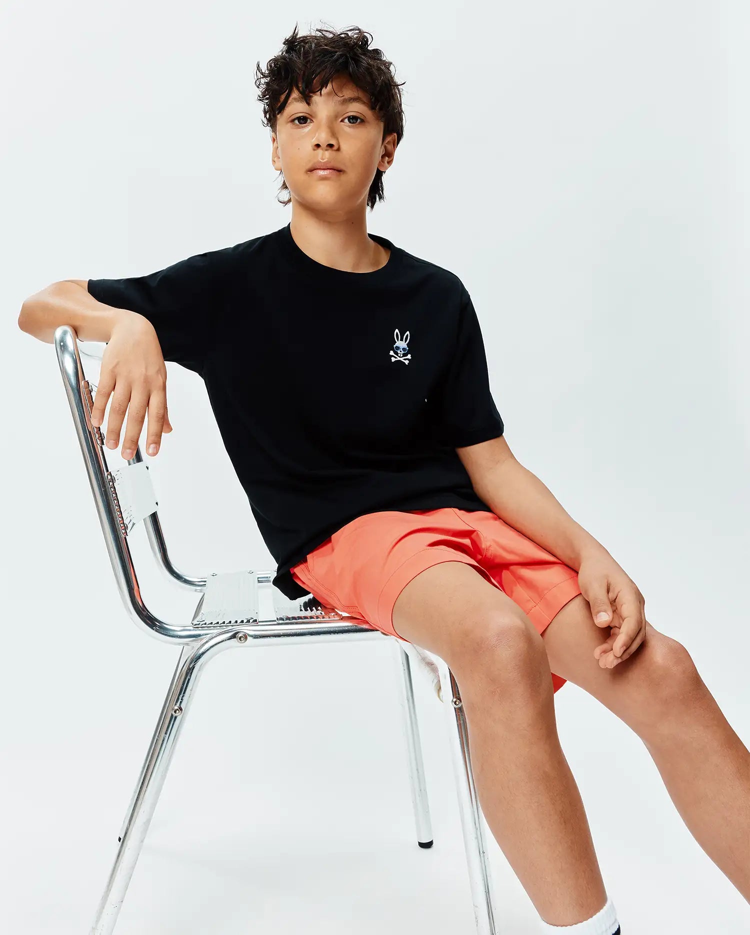 Black Kids Sunny Chest Graphic Tee in smooth jersey knit, featuring a small embroidered white and blue Psycho Bunny skull logo on the left chest, paired with coral shorts.