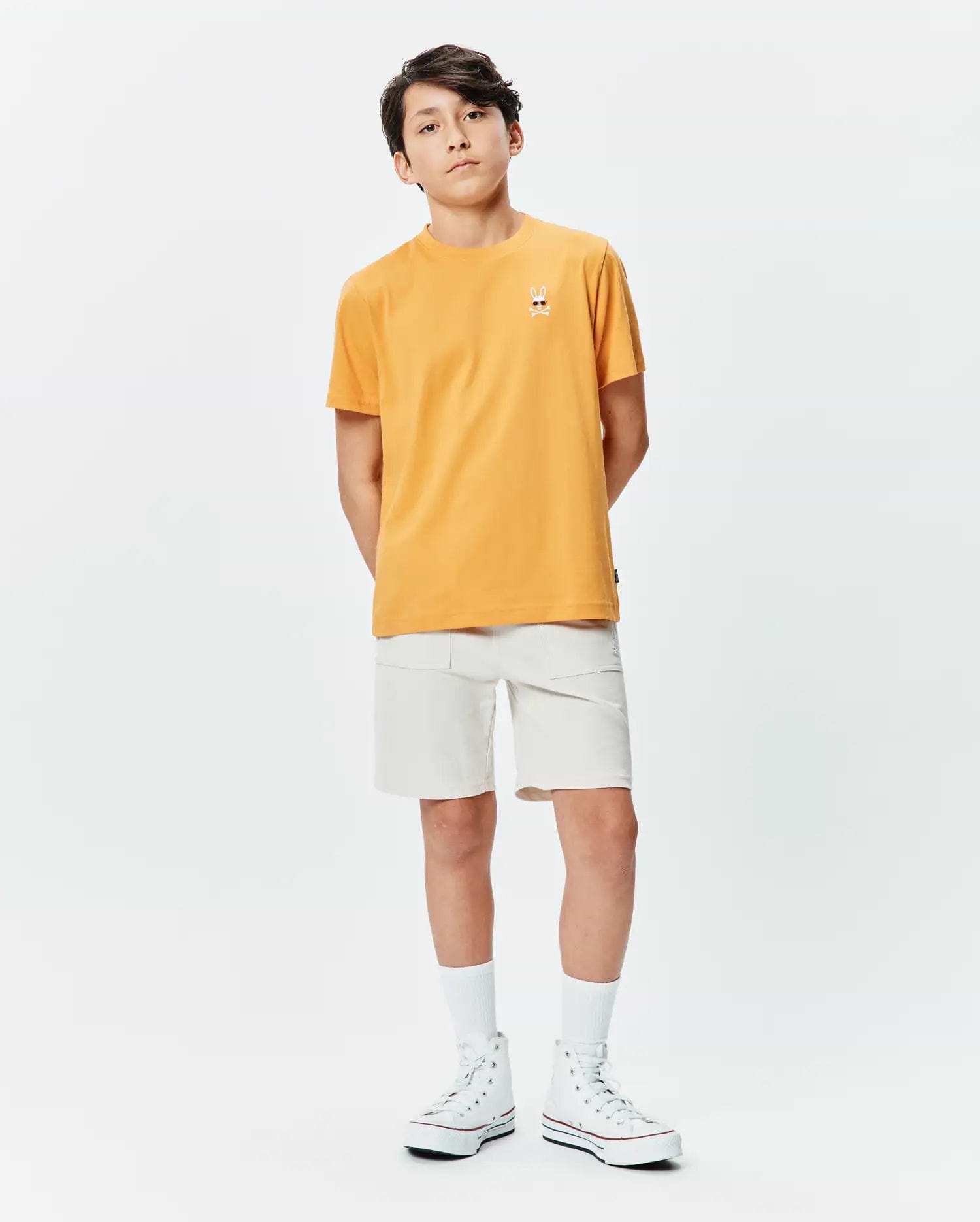 Orange Kids Sunny Chest Graphic Tee in classic fit jersey, featuring a small embroidered Psycho Bunny skull logo on the left chest, paired with off-white woven shorts.
