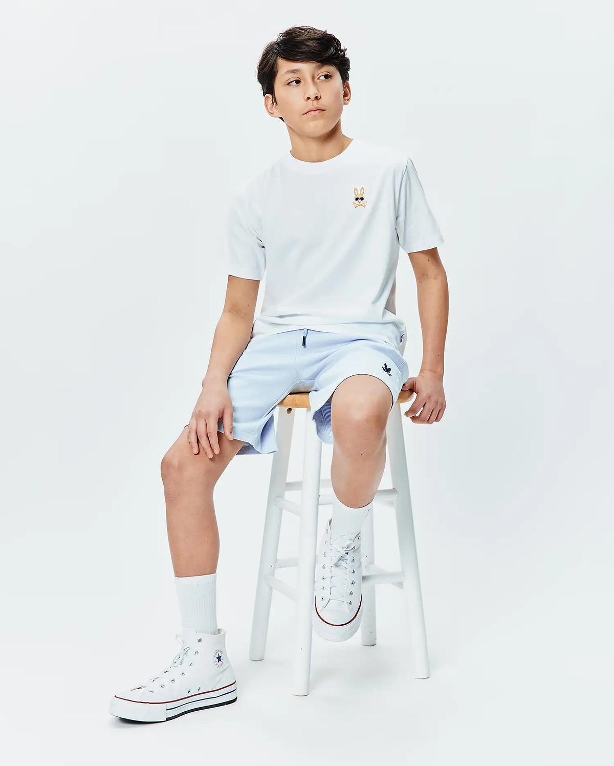 Kids Sunny Chest Graphic Tee, a white jersey t-shirt with a small embroidered Psycho Bunny skull in sunglasses on the left chest, paired with light blue relaxed-fit shorts featuring a small embroidered bunny logo on the right thigh.