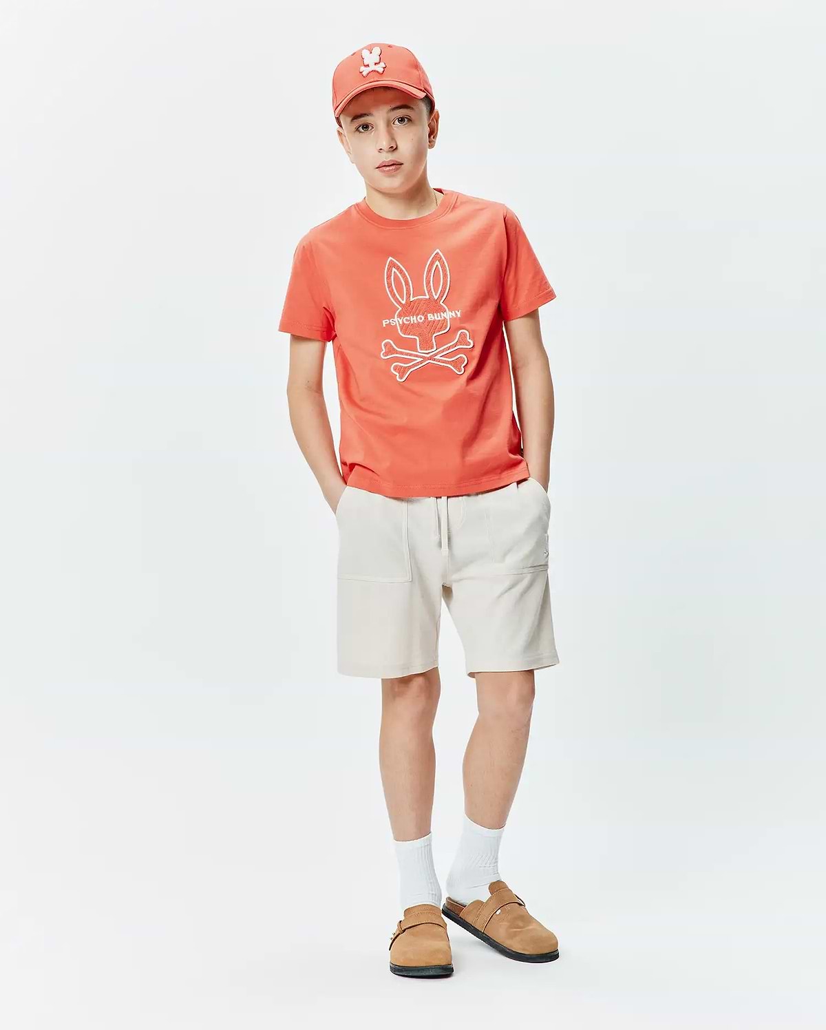 Kids Craig Graphic Tee, a classic fit coral jersey t-shirt with a prominent white Psycho Bunny skull and crossbones graphic on the center chest.