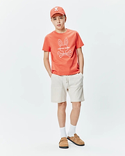 Kids Craig Graphic Tee, a classic fit coral jersey t-shirt with a prominent white Psycho Bunny skull and crossbones graphic on the center chest.