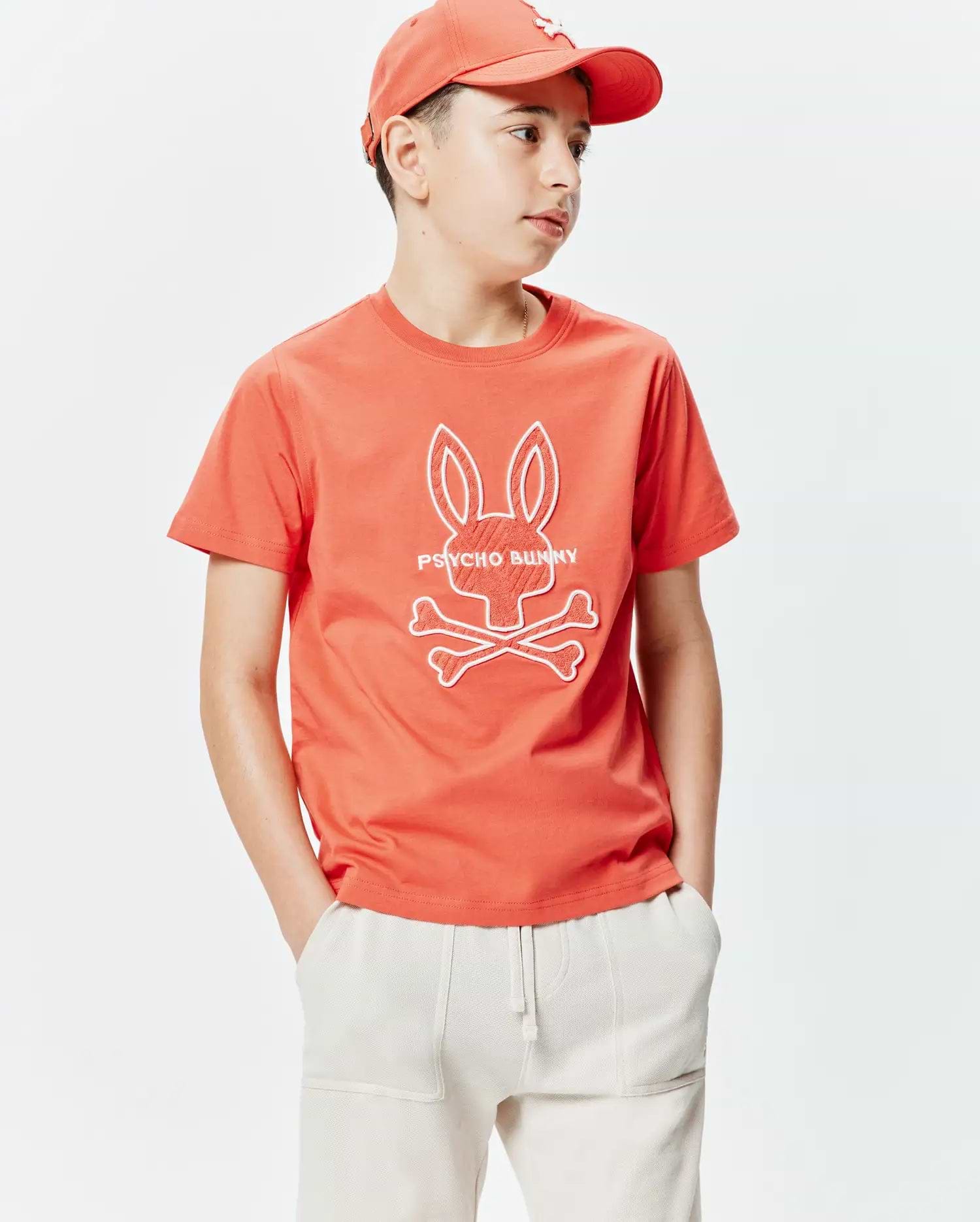 Kids Craig Graphic Tee in coral orange, featuring a classic fit and soft jersey knit. A large, textured white Psycho Bunny skull and crossbones graphic with PSYCHO BUNNY text is centered on the chest.
