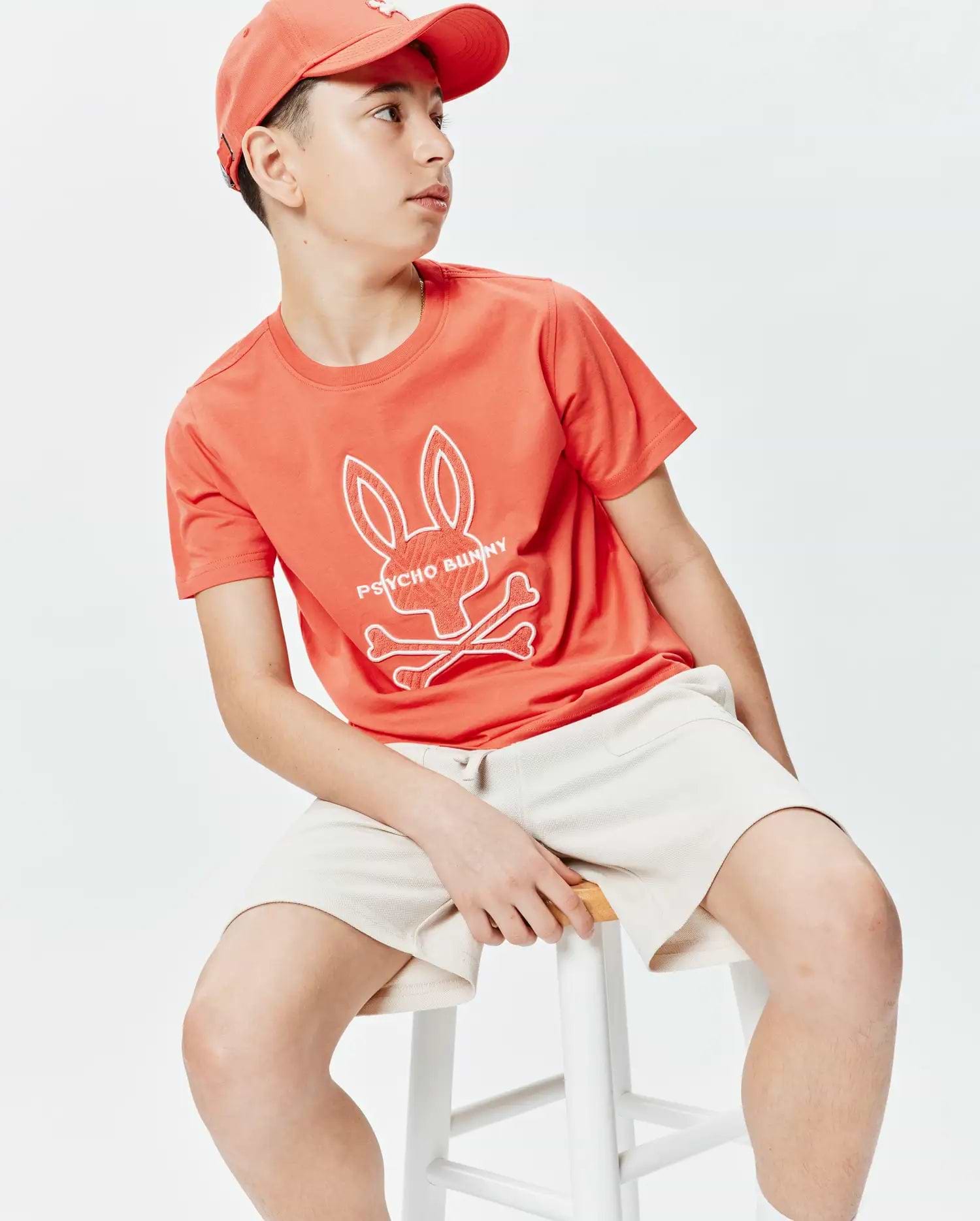 Kids Craig Graphic Tee, a coral classic-fit jersey t-shirt with a large white Psycho Bunny skull and crossbones chest graphic, paired with cream textured knit shorts and a coral baseball cap.