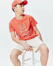 Kids Craig Graphic Tee, a coral classic-fit jersey t-shirt with a large white Psycho Bunny skull and crossbones chest graphic, paired with cream textured knit shorts and a coral baseball cap.