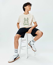 Kids Craig Graphic Tee, a cream jersey t-shirt with a large green Psycho Bunny skull and crossbones logo on the chest, paired with classic fit black shorts. (4)