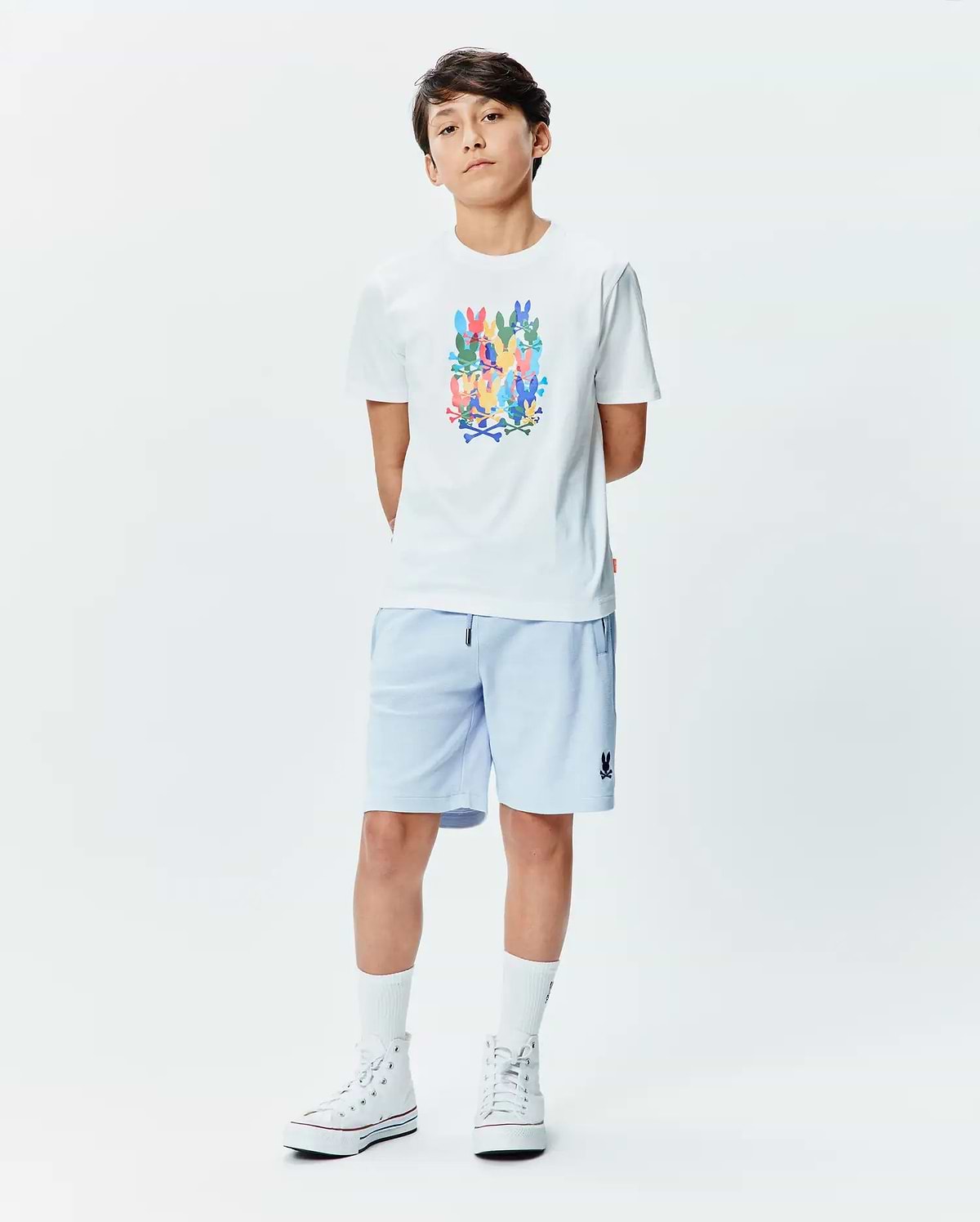 White Kids Tanner Graphic Tee with a large multicolor stacked Psycho Bunny skull logo print on the chest, paired with light blue relaxed-fit shorts featuring a small embroidered dark blue Psycho Bunny logo on the left leg.