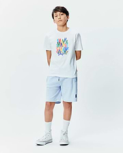 White Kids Tanner Graphic Tee with a large multicolor stacked Psycho Bunny skull logo print on the chest, paired with light blue relaxed-fit shorts featuring a small embroidered dark blue Psycho Bunny logo on the left leg.