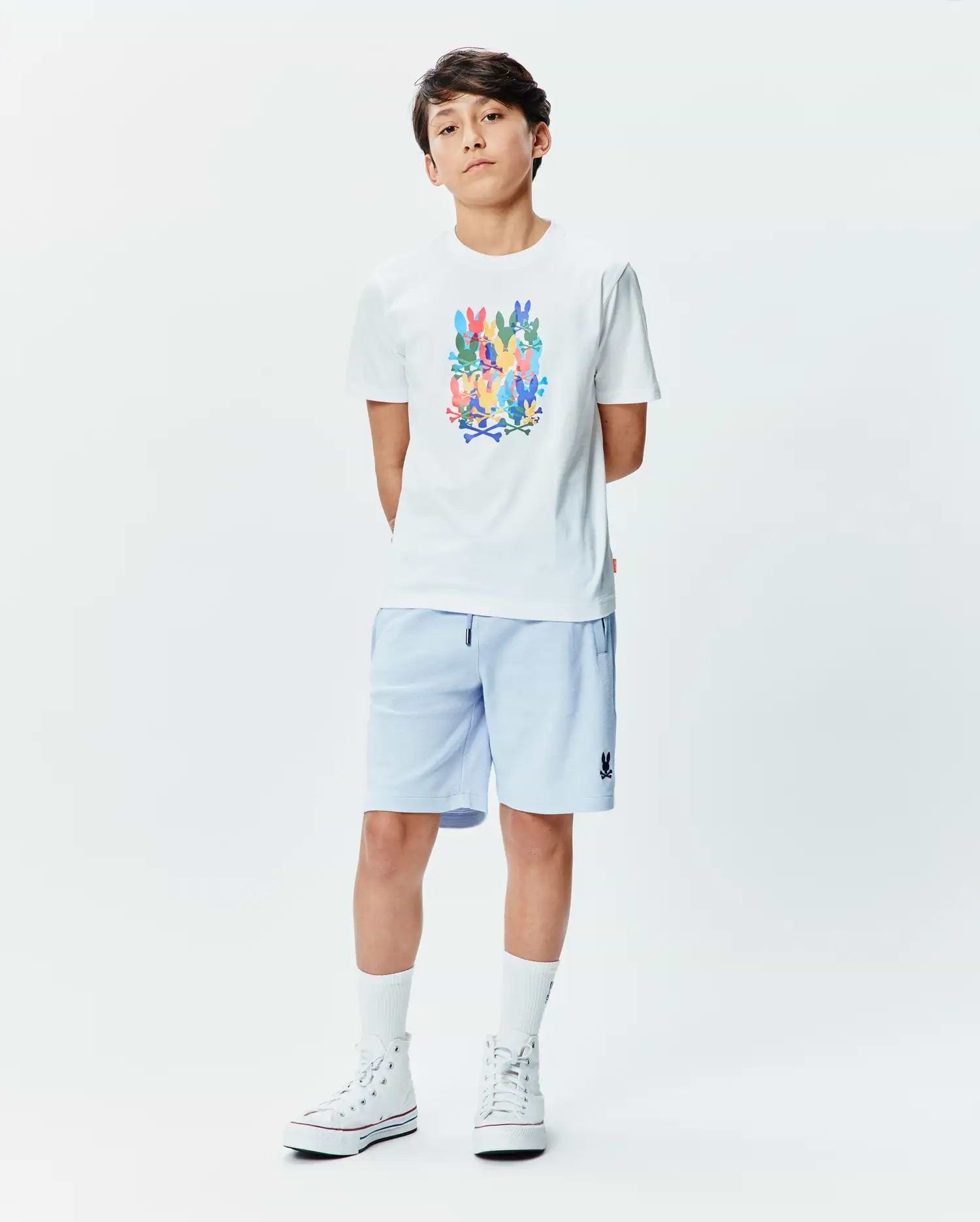 White Kids Tanner Graphic Tee with a large multicolor stacked Psycho Bunny skull logo print on the chest, paired with light blue relaxed-fit shorts featuring a small embroidered dark blue Psycho Bunny logo on the left leg.