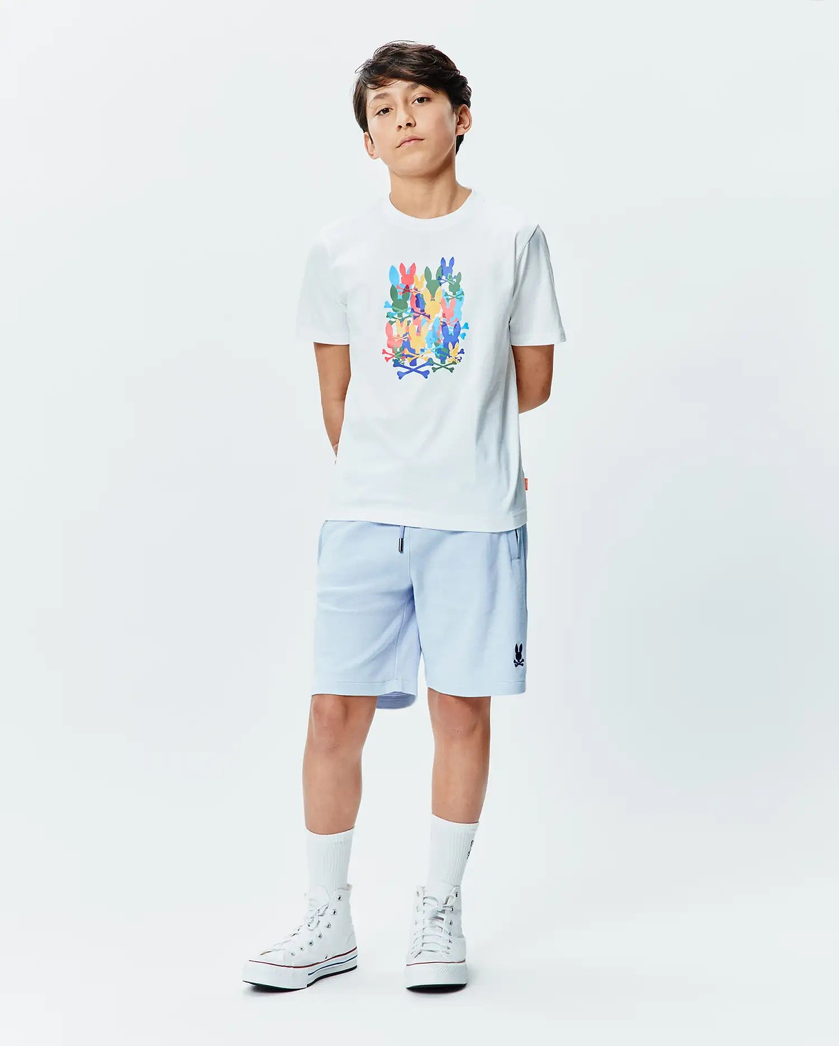 White Kids Tanner Graphic Tee with a large multicolor stacked Psycho Bunny skull logo print on the chest, paired with light blue relaxed-fit shorts featuring a small embroidered dark blue Psycho Bunny logo on the left leg.
