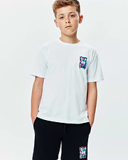 Kids Isaac Relaxed Fit Tee, a white jersey t-shirt featuring a vibrant, abstract Psycho Bunny skull logo graphic on the left chest. Black shorts with a matching colorful bunny skull logo on the left thigh complete the look.
