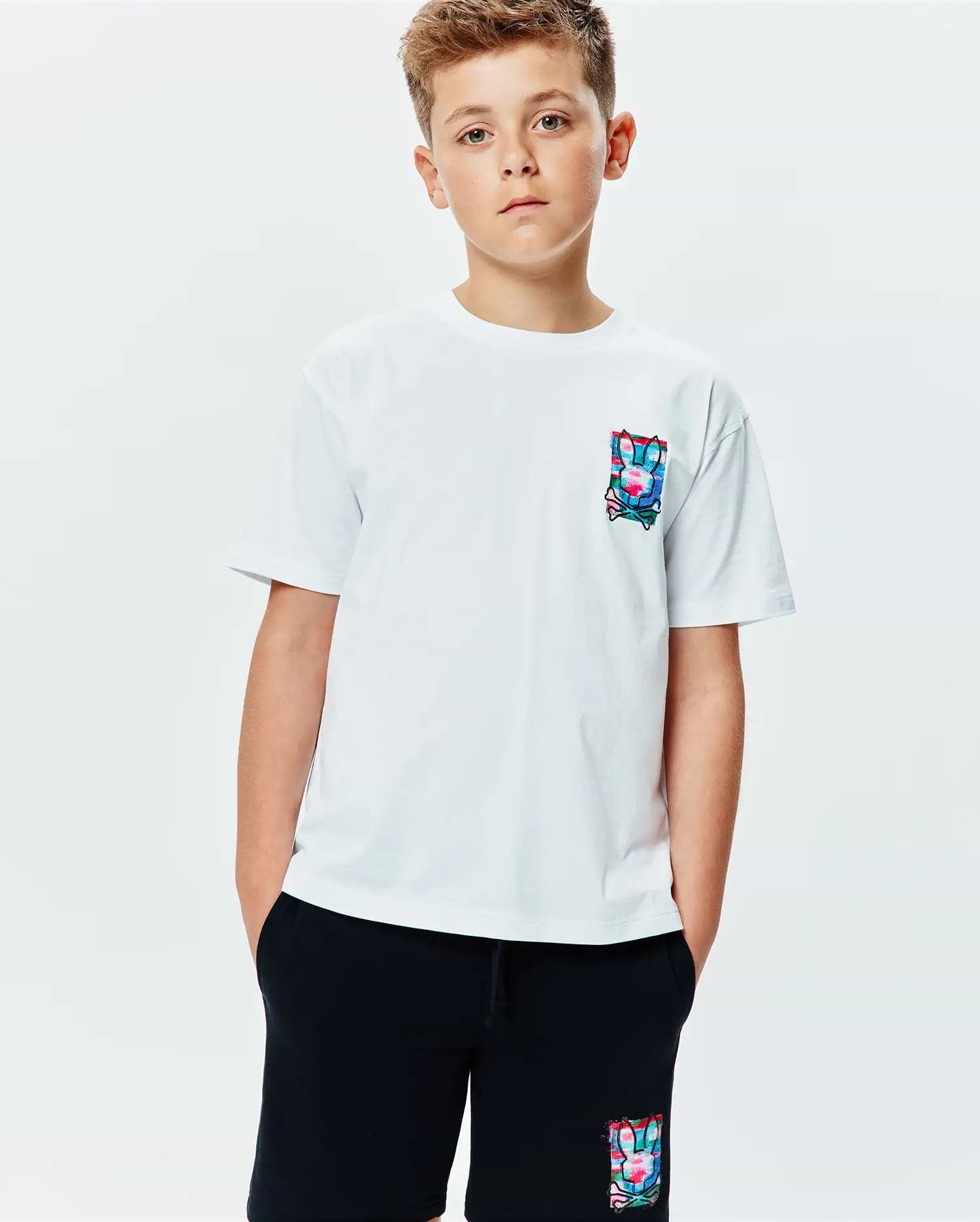 Kids Isaac Relaxed Fit Tee, a white jersey t-shirt featuring a vibrant, abstract Psycho Bunny skull logo graphic on the left chest. Black shorts with a matching colorful bunny skull logo on the left thigh complete the look.