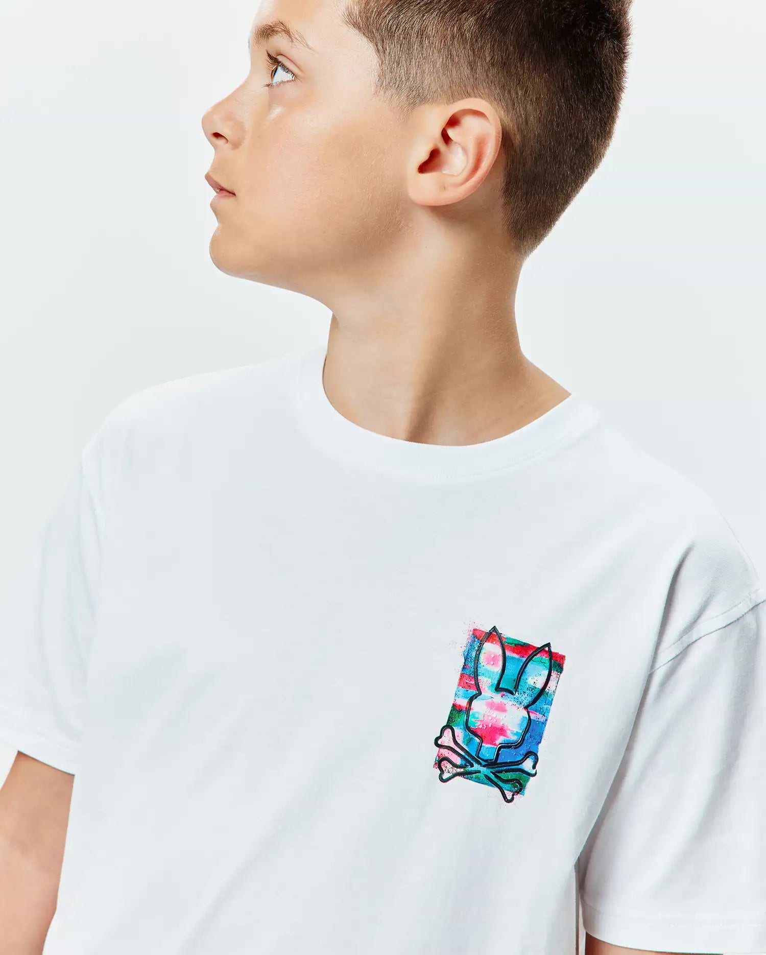 Kids Isaac Relaxed Fit Tee, a white relaxed fit jersey t-shirt with a small chest graphic featuring the iconic Psycho Bunny skull logo over a vibrant blue, pink, and green distressed background.
