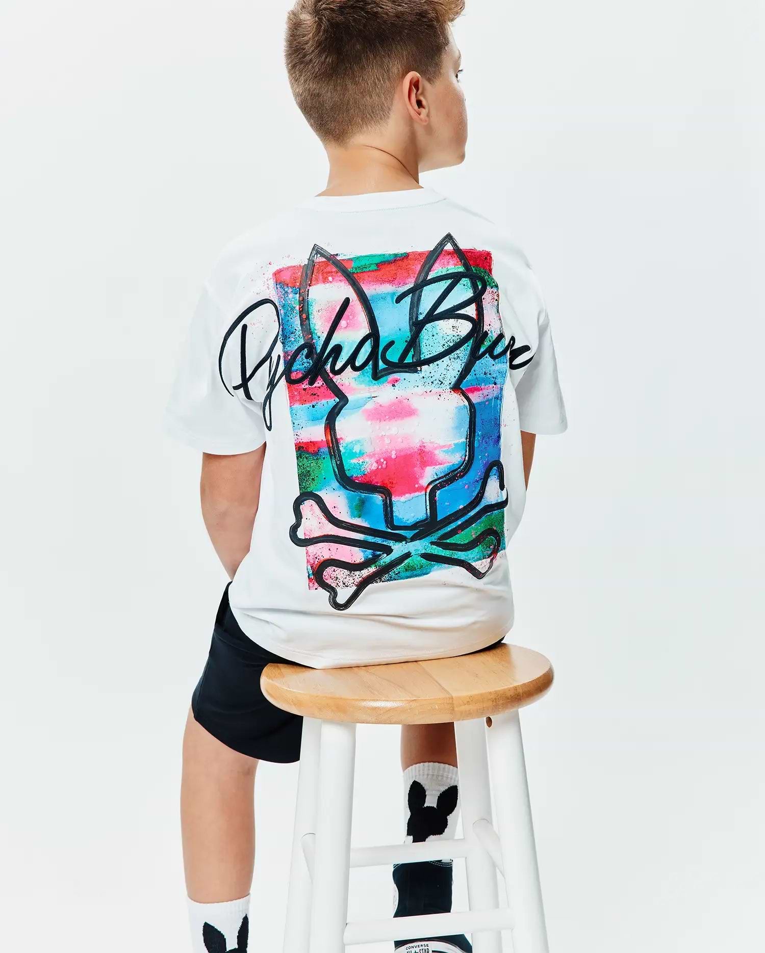 Kids Isaac Relaxed Fit Tee, white jersey t-shirt with a large back graphic featuring the Psycho Bunny skull and crossbones over a vibrant multi-color paint-splatter design and black script logo.